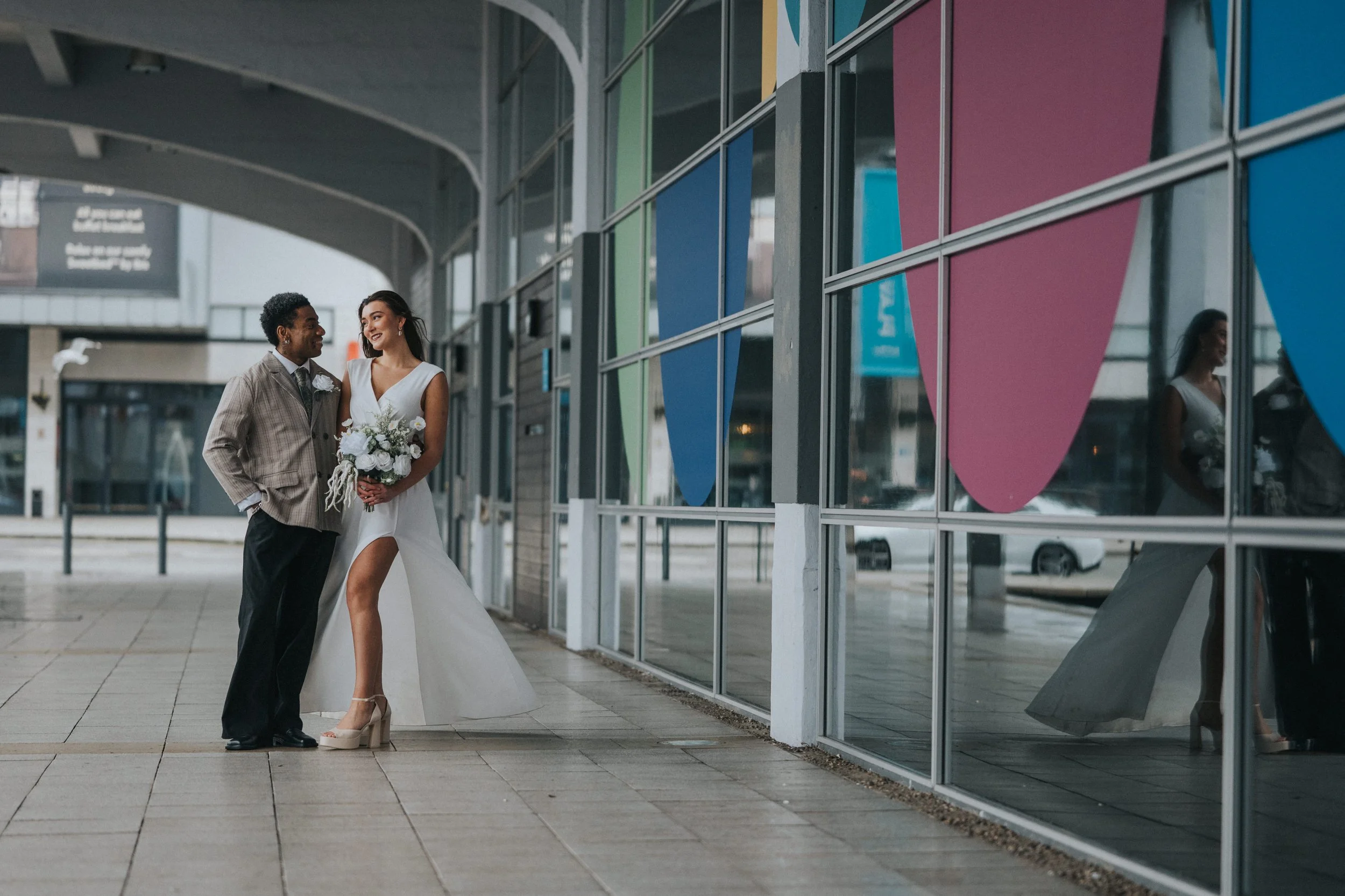 A couple dressed in wedding attire standing outside a modern building with colorful window panels, smiling and looking at each other.