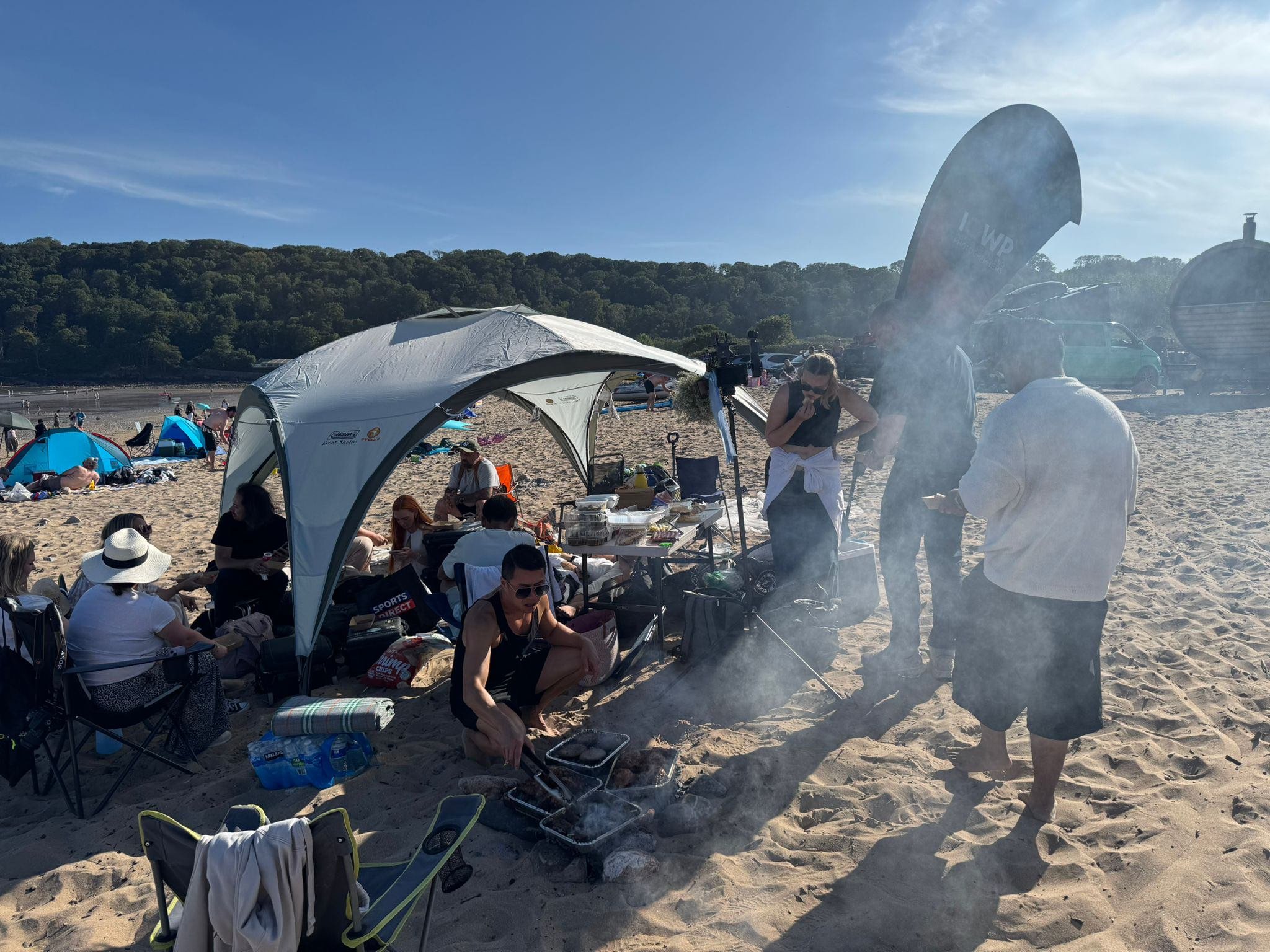 People gathered on a sandy beach under a canopy, some sitting and some standing, with a barbecue grill and various beach gear. In the background, there are more beachgoers, umbrellas, and boats on the water, with a hill covered in trees and a blue sk