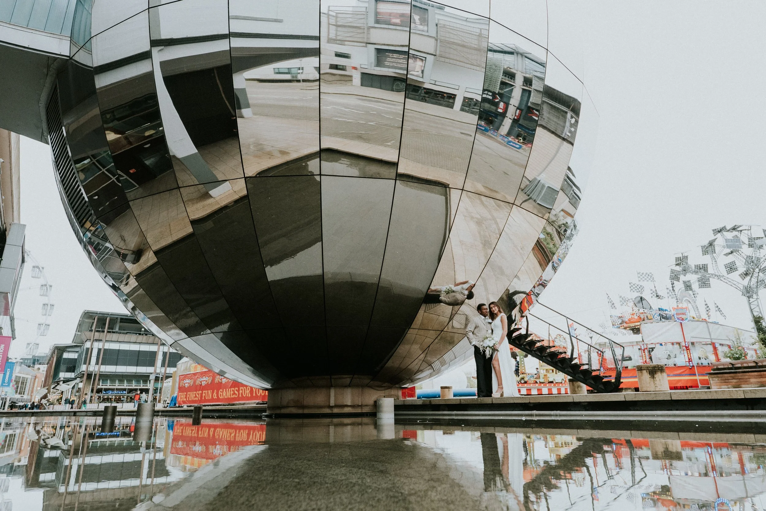 Reflection of a modern building with a unique, curved, shiny facade, two people taking a photo in front, cityscape with signs for entertainment and shops in the background.