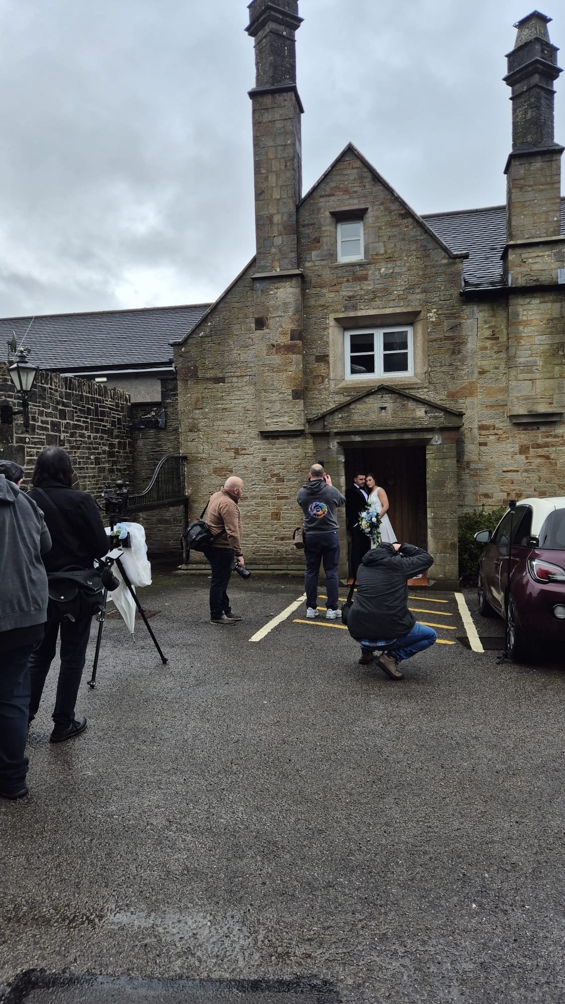 Couple in wedding attire taking photos outside a historic stone building with photographers and onlookers around.
