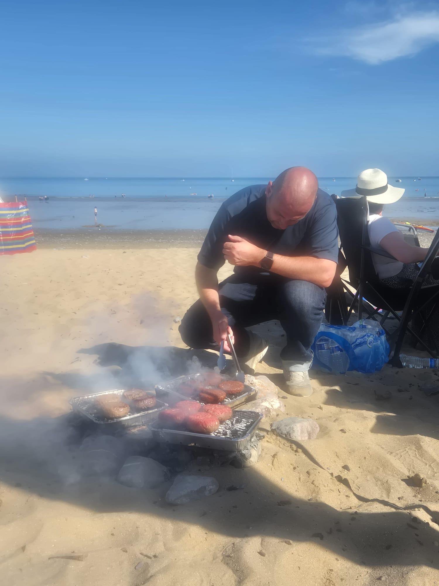 A man is grilling hamburgers on a barbecue grill on a sandy beach, with the ocean and a clear blue sky in the background. There is a woman sitting nearby wearing a hat and sunglasses, and some chairs and water bottles around them.