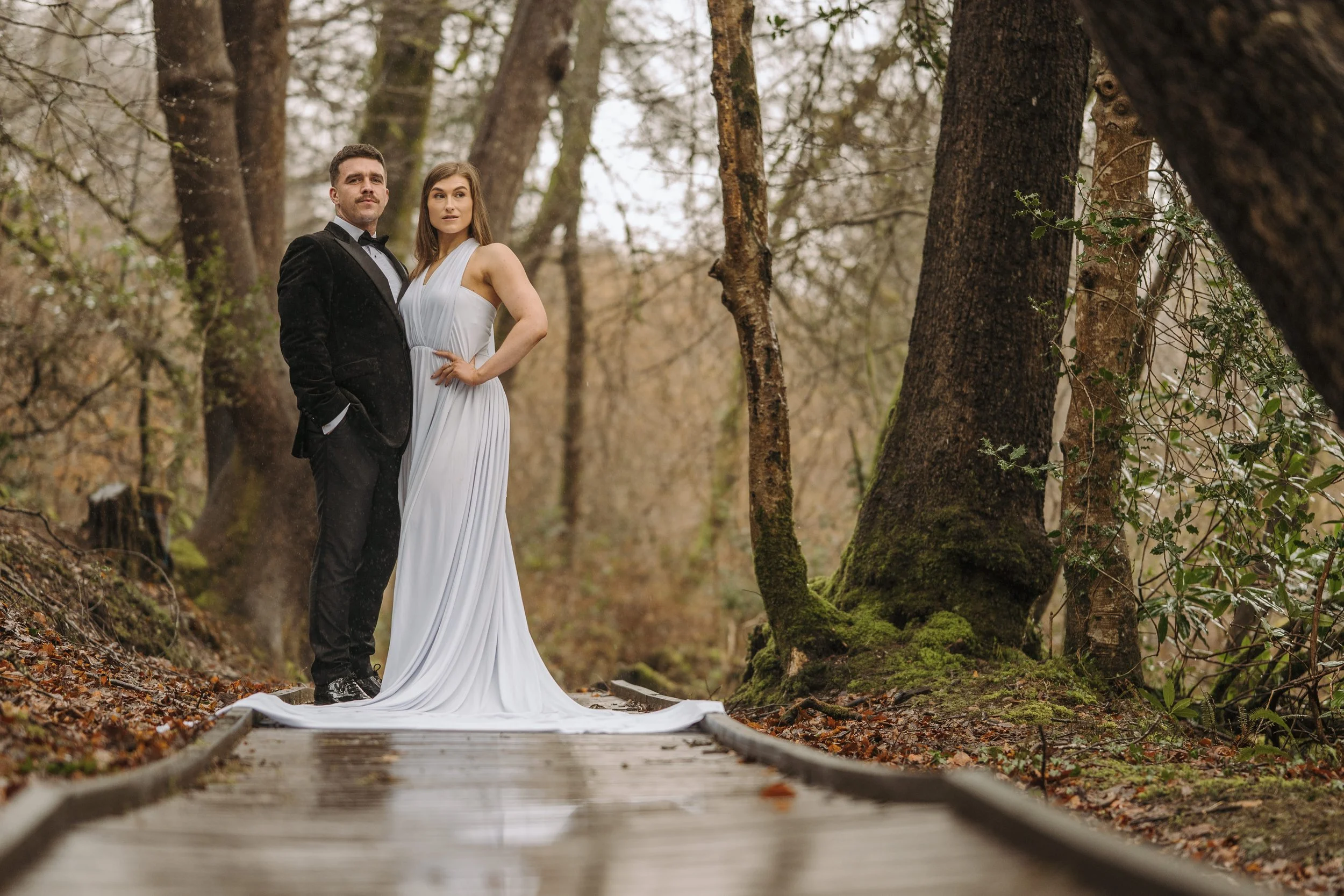 A bride and groom standing on a wooden pathway in a forest, dressed in formal wedding attire, with the bride in a white gown and the groom in a black tuxedo.