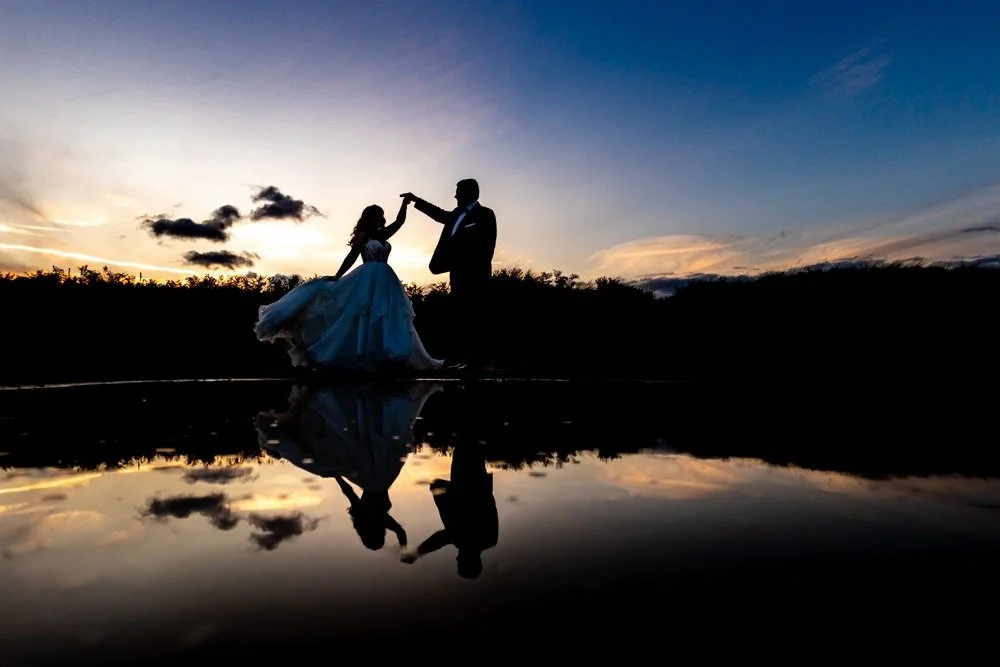 Silhouettes of a man and woman dancing on a hill at sunset, with their reflections visible in a body of water below.