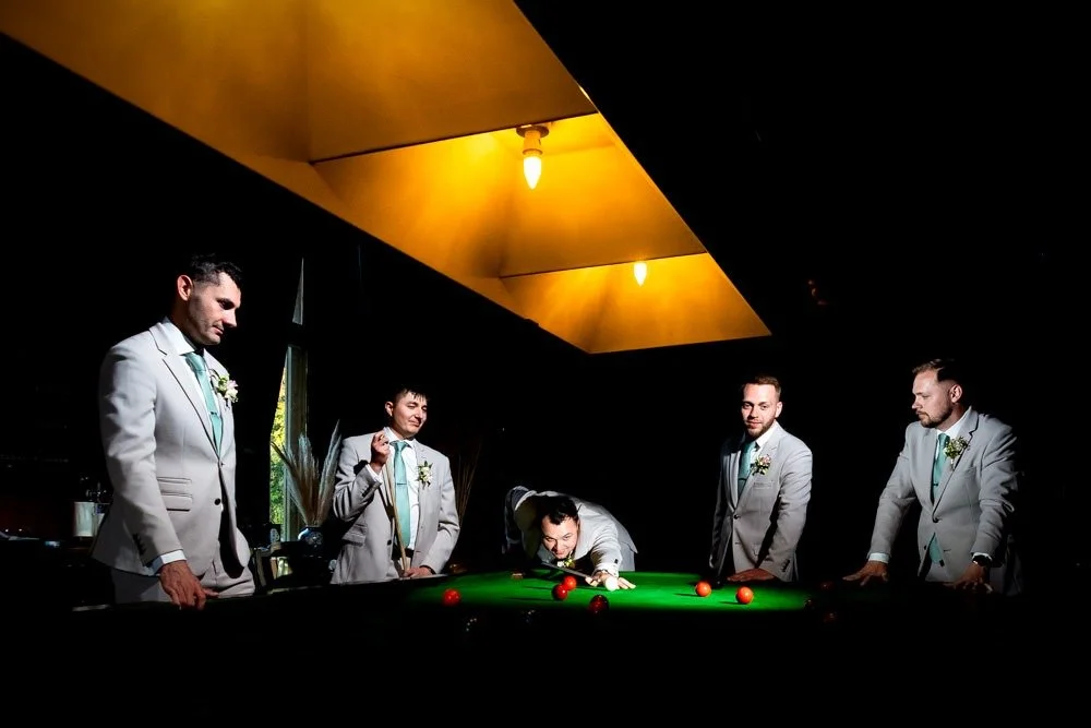 Groom and groomsmen in tuxedos playing snooker or pool in a dark room with yellow ceiling lights.