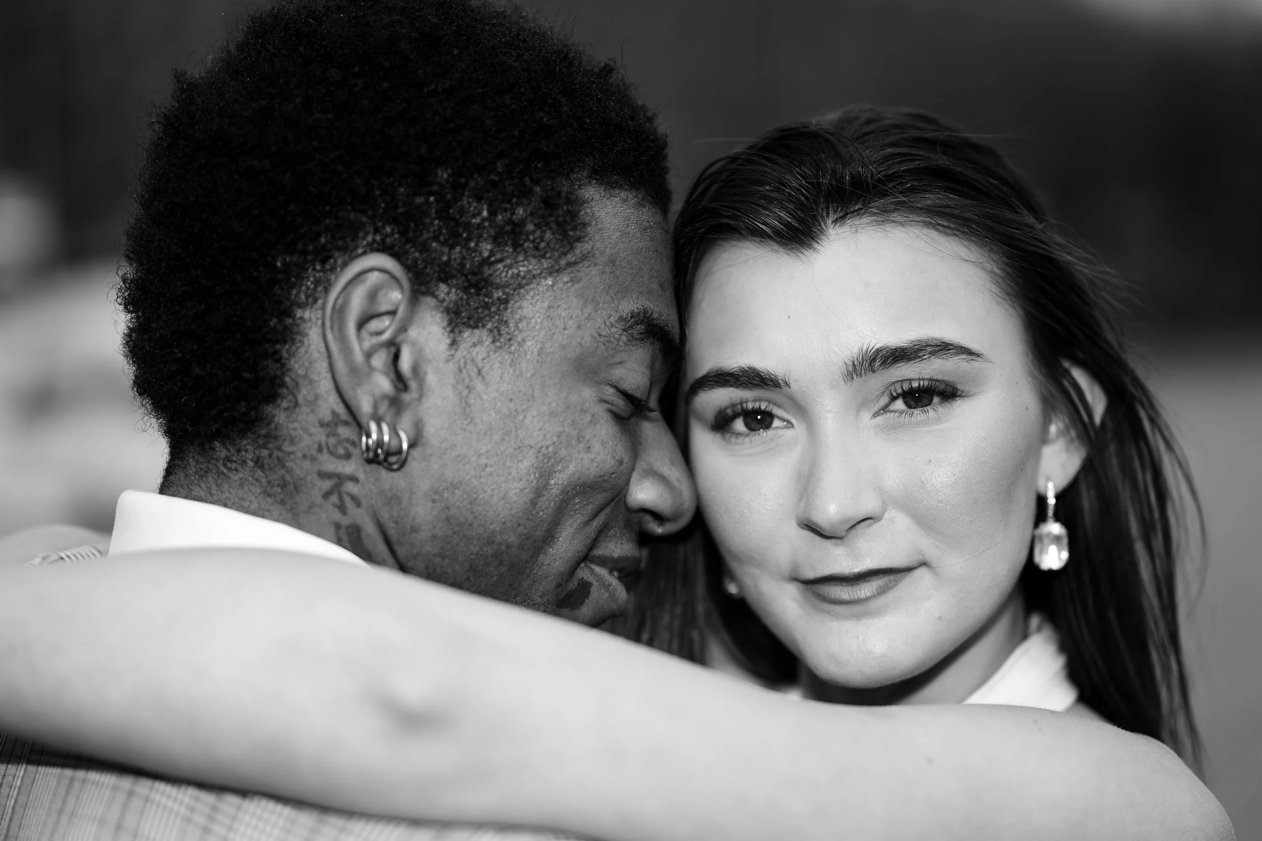 A black-and-white photo of an interracial couple embracing closely, with the man's face pressed against the woman's to show affection, and the woman smiling gently while looking at the camera.