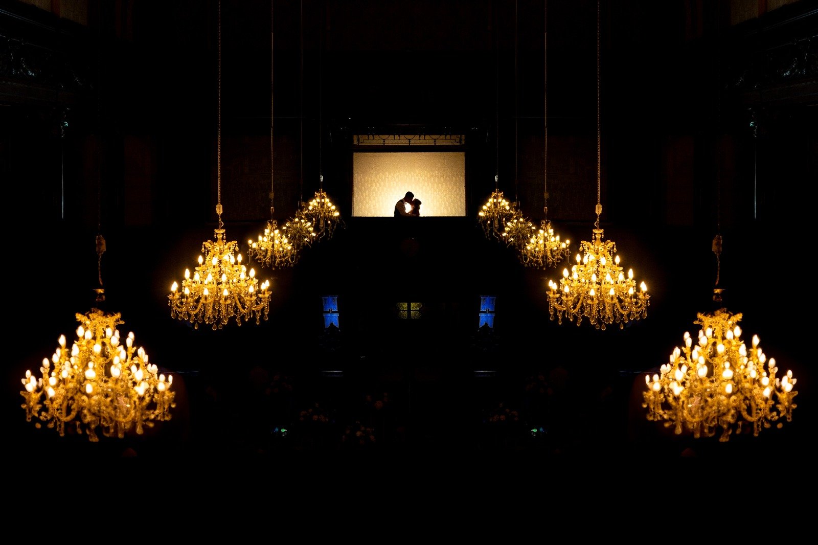 Silhouette of a couple kissing on a balcony at the top of a darkened room with multiple chandeliers hanging from the ceiling, illuminating the space with warm light.