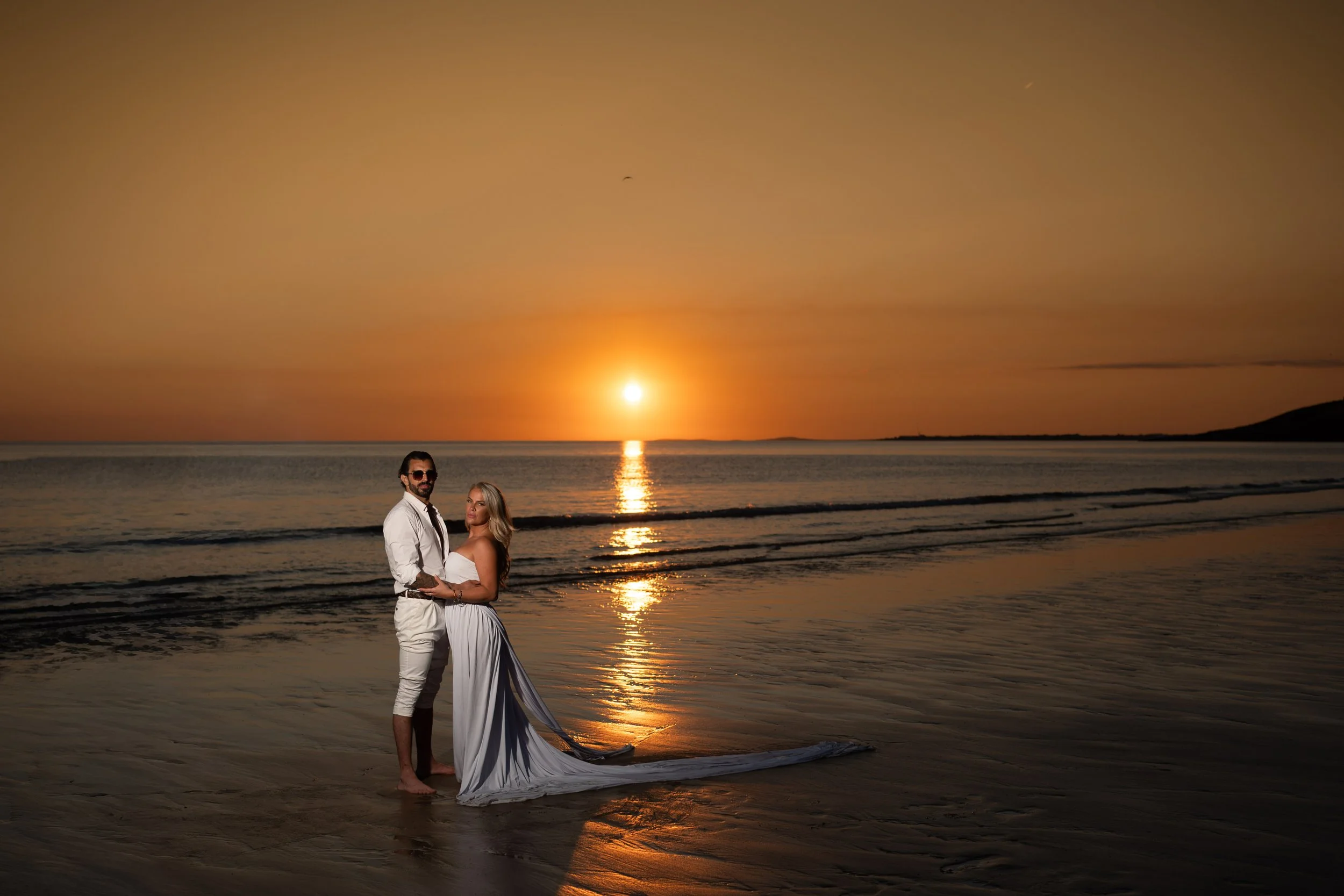 A couple dressed in wedding attire standing on the beach during sunset, with the man wearing a white shirt and sunglasses and the woman in a white strapless dress with a long train, holding each other as the reflection of the setting sun shines on th