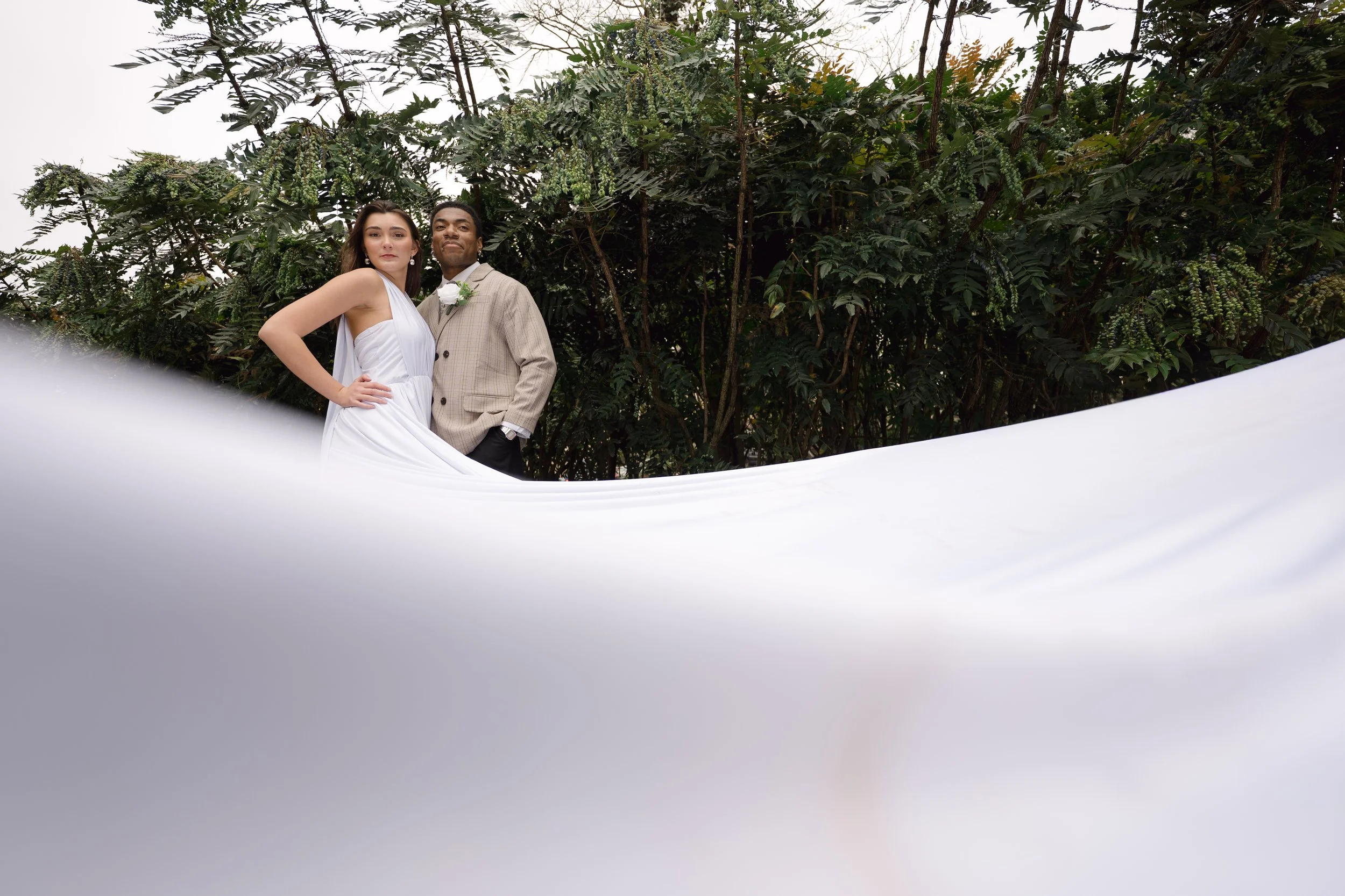 A couple dressed in wedding attire, standing outdoors in front of dense green foliage, with a white fabric covering part of the foreground.