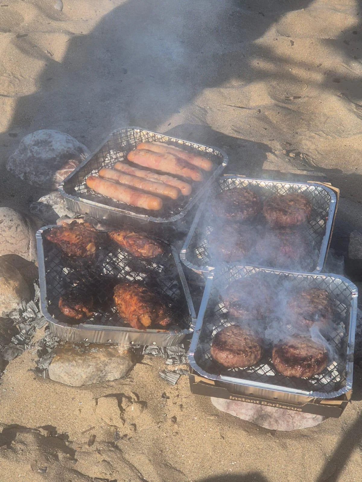 Aluminum foil trays with hotdogs, chicken, and hamburgers grilling on a beach fire over rocks.