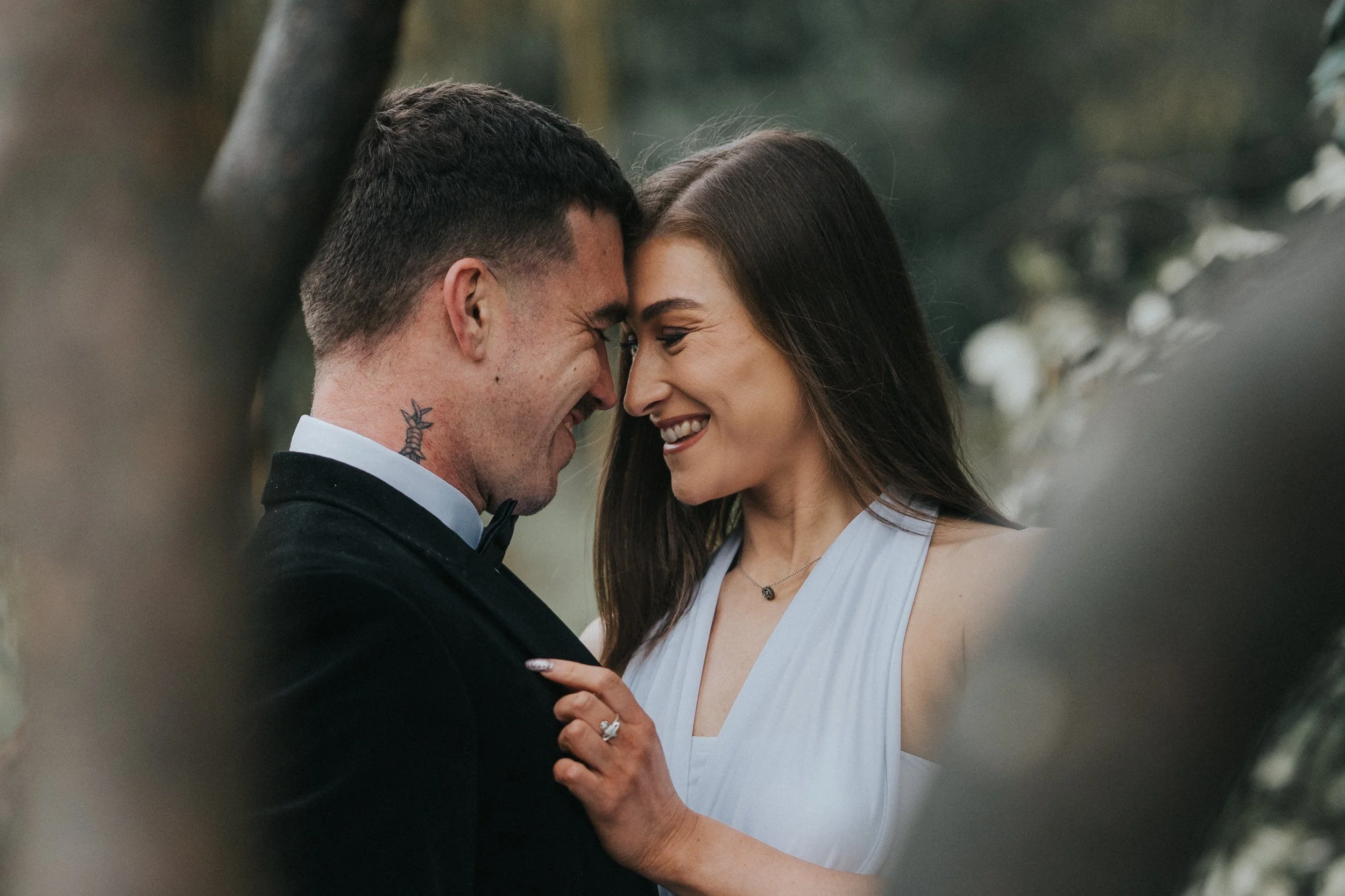 A couple in formal attire, smiling and touching foreheads, outdoors amongst trees.