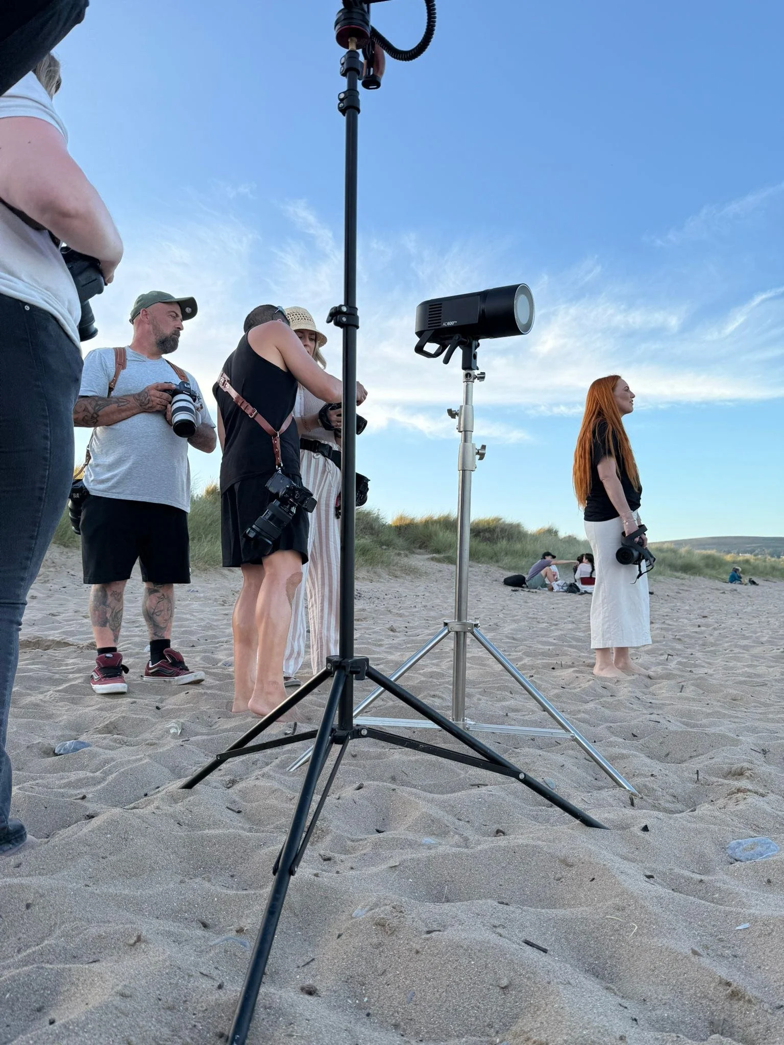 Group of people on the beach with photography equipment, including a woman standing barefoot holding a camera, surrounded by others with cameras and lenses, under a partly cloudy sky.