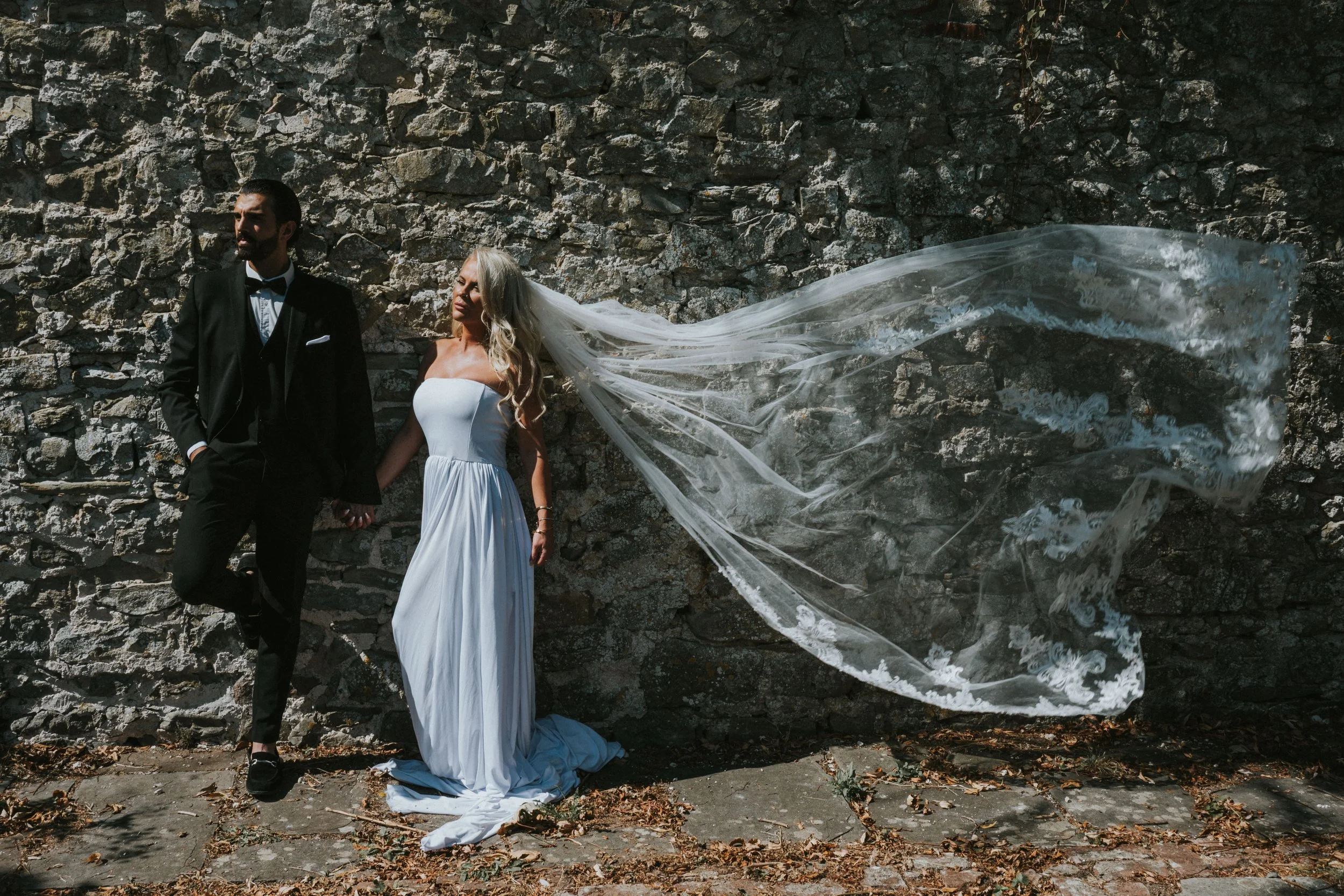 A newlywed couple stands in front of a stone wall, holding hands. The groom wears a black tuxedo and the bride wears a white wedding gown with a long, flowing veil.