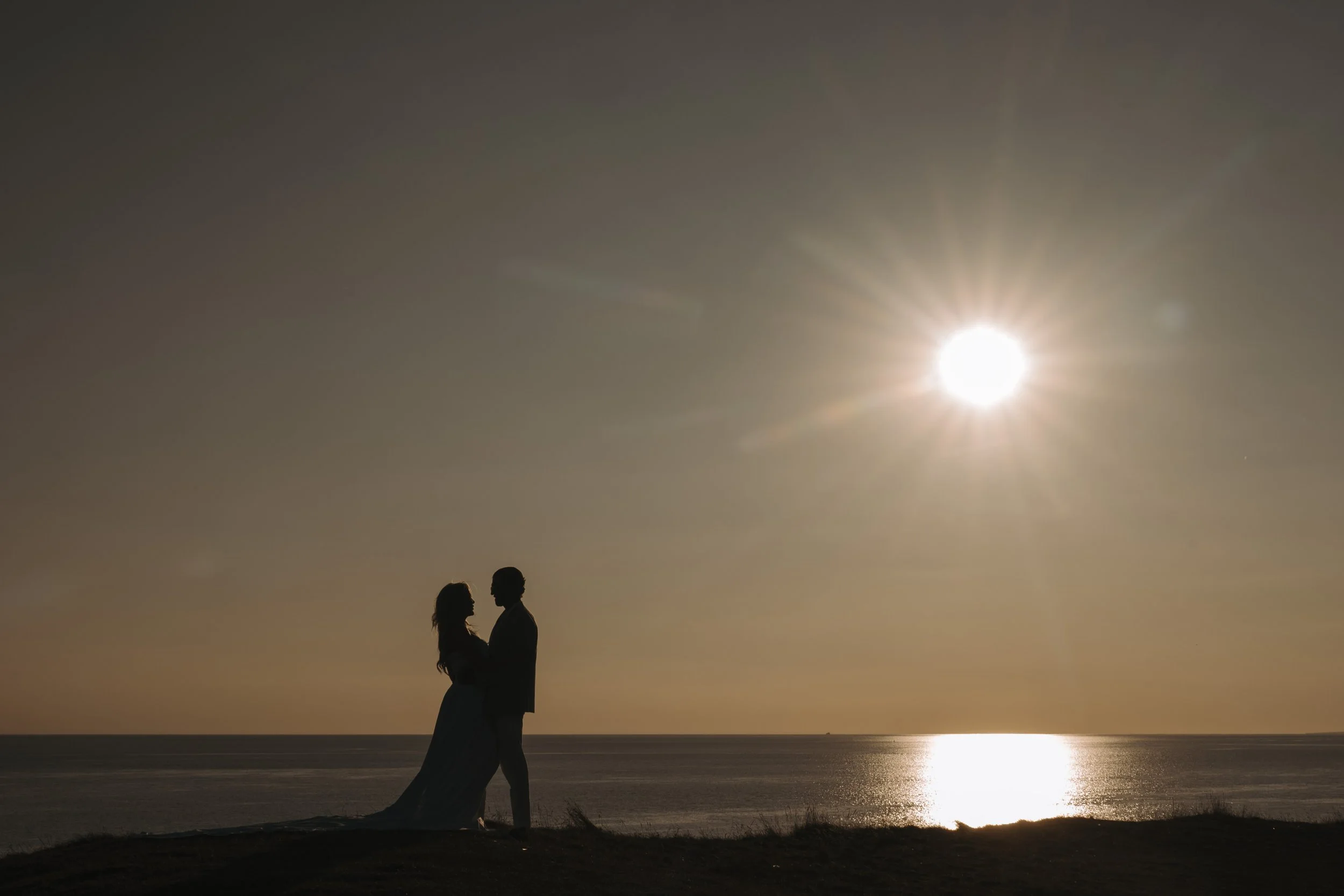 Silhouetted couple standing close together on beach at sunset with the sun low in the sky and reflecting on water.