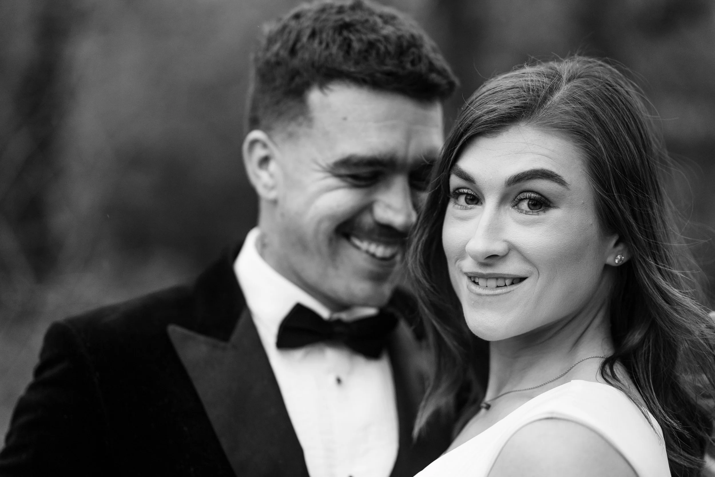 Black and white photo of a smiling young couple dressed in formal attire, with the man wearing a tuxedo and the woman wearing a dress, outdoors.