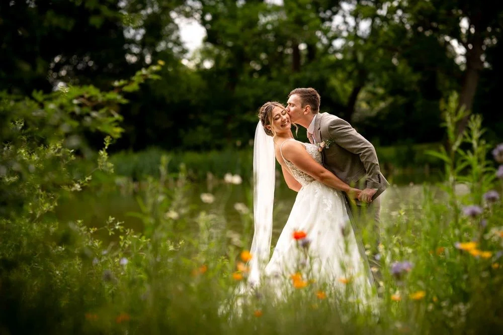 A smiling bride and groom embrace outdoors in a lush, green setting, with a pond and trees in the background, during their wedding photos.