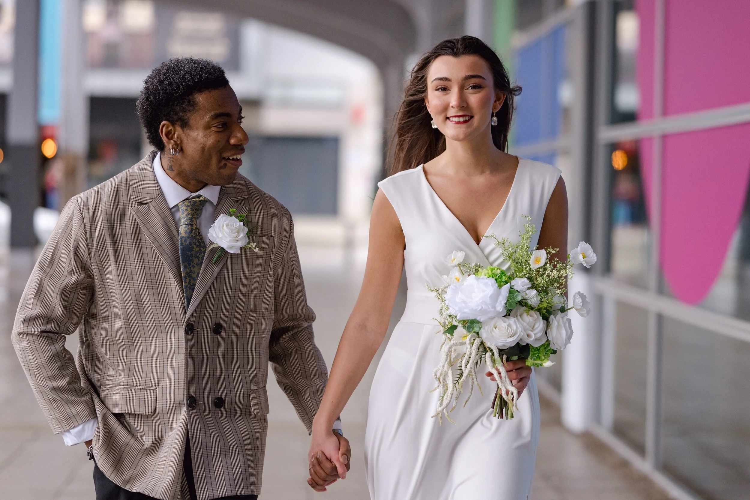 A smiling woman in a white wedding dress holding a bouquet of white flowers walks hand in hand with a man in a plaid suit with a boutonniere in an indoor setting with colorful walls.