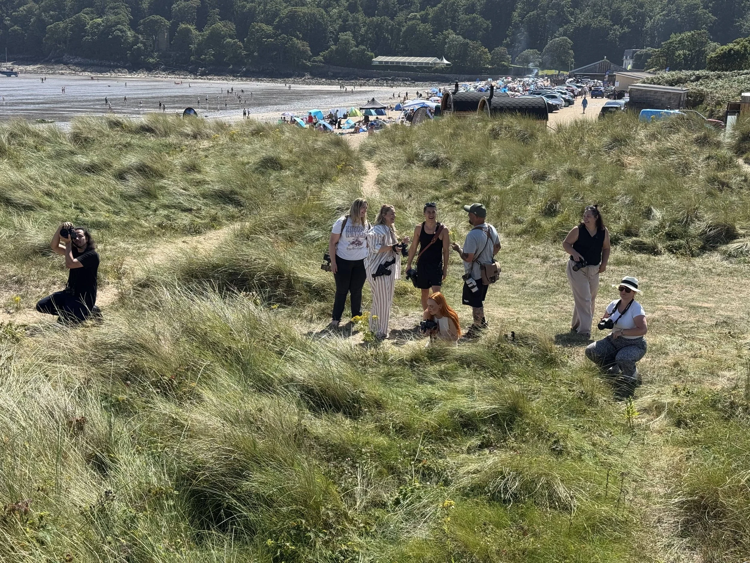 A group of people posing for photographs in grassy dunes near a beach, with many beachgoers and umbrellas in the background.