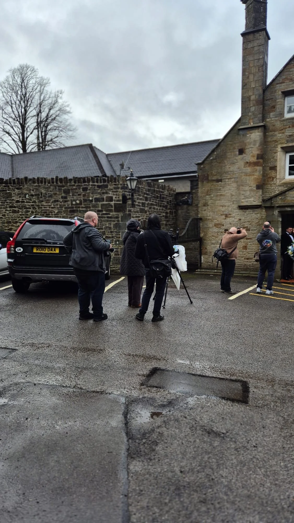 A group of people standing outside a stone building on a cloudy day, some taking photos or looking towards the building, with cars parked nearby on an asphalt lot.