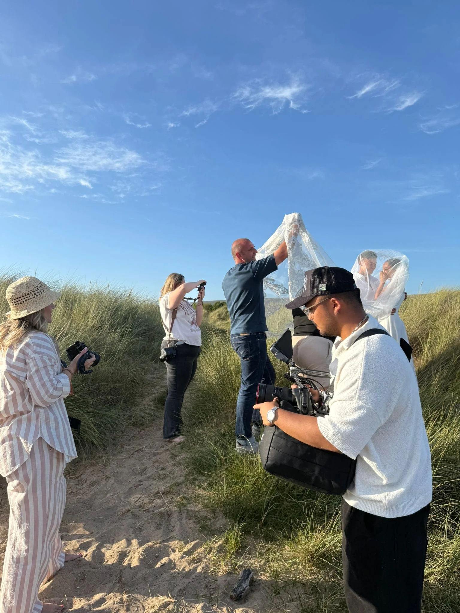 A group of people, including a bride and groom under a veil, on a sandy grassy path outdoors with blue sky in the background. Some are taking photos or videos while a man helps with the veil.