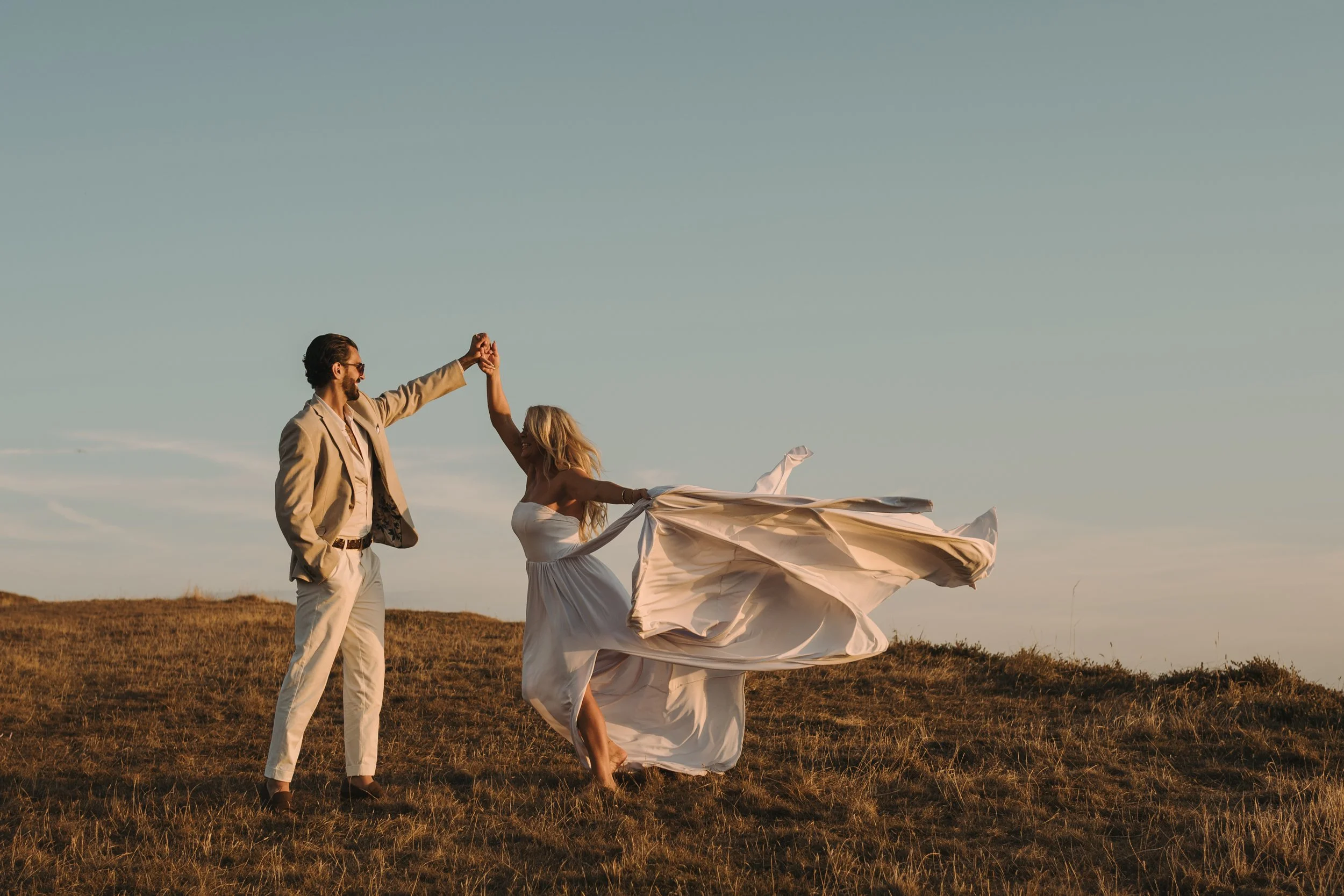 A man and woman dancing outdoors in a field during sunset, with the woman twirling in a flowing white dress and the man in a beige suit holding her hand.
