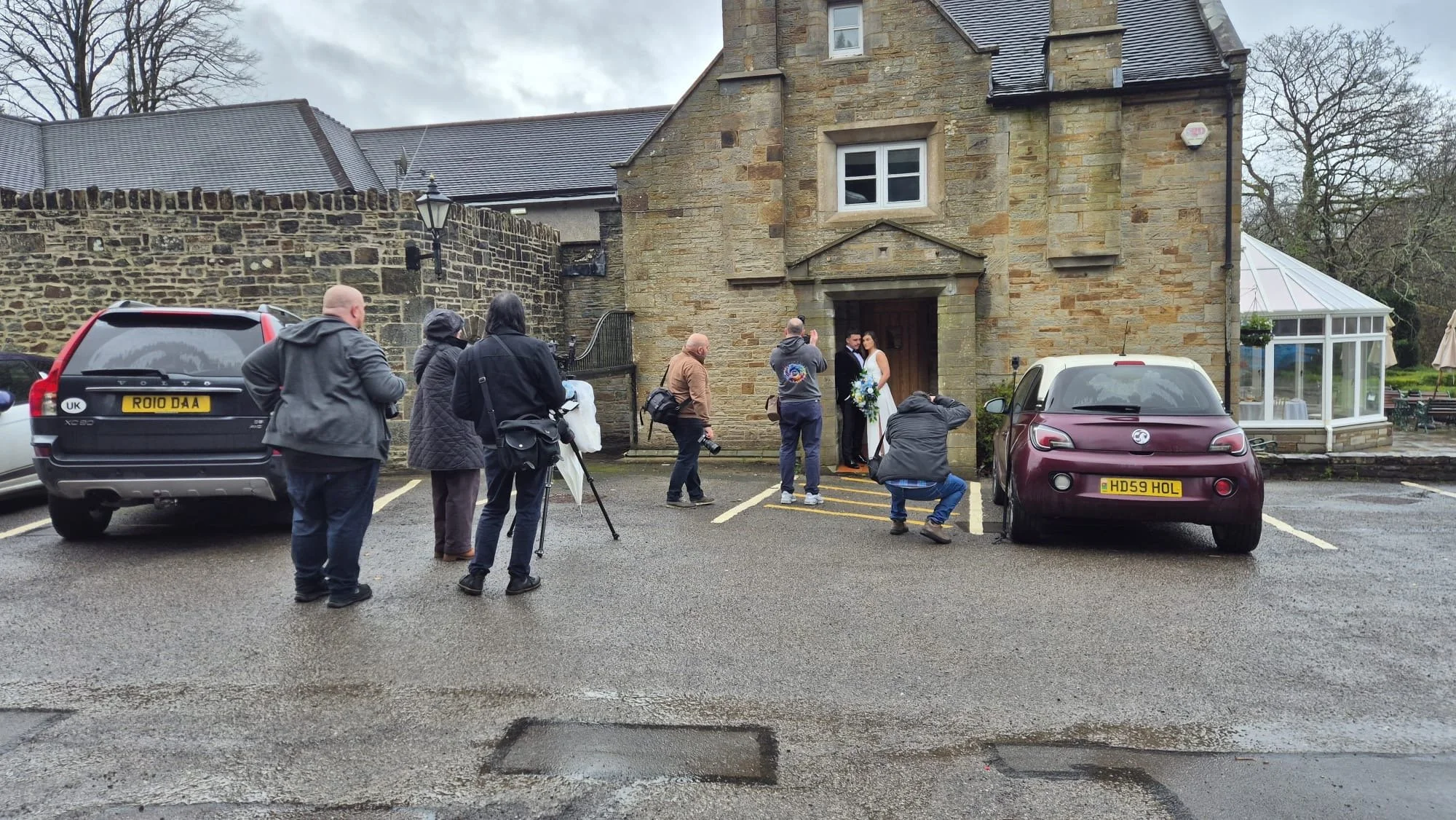 A wedding ceremony outside a stone building with a bride and groom standing at the entrance, surrounded by photographers and guests. Cars are parked in front of the building, and the sky is overcast.
