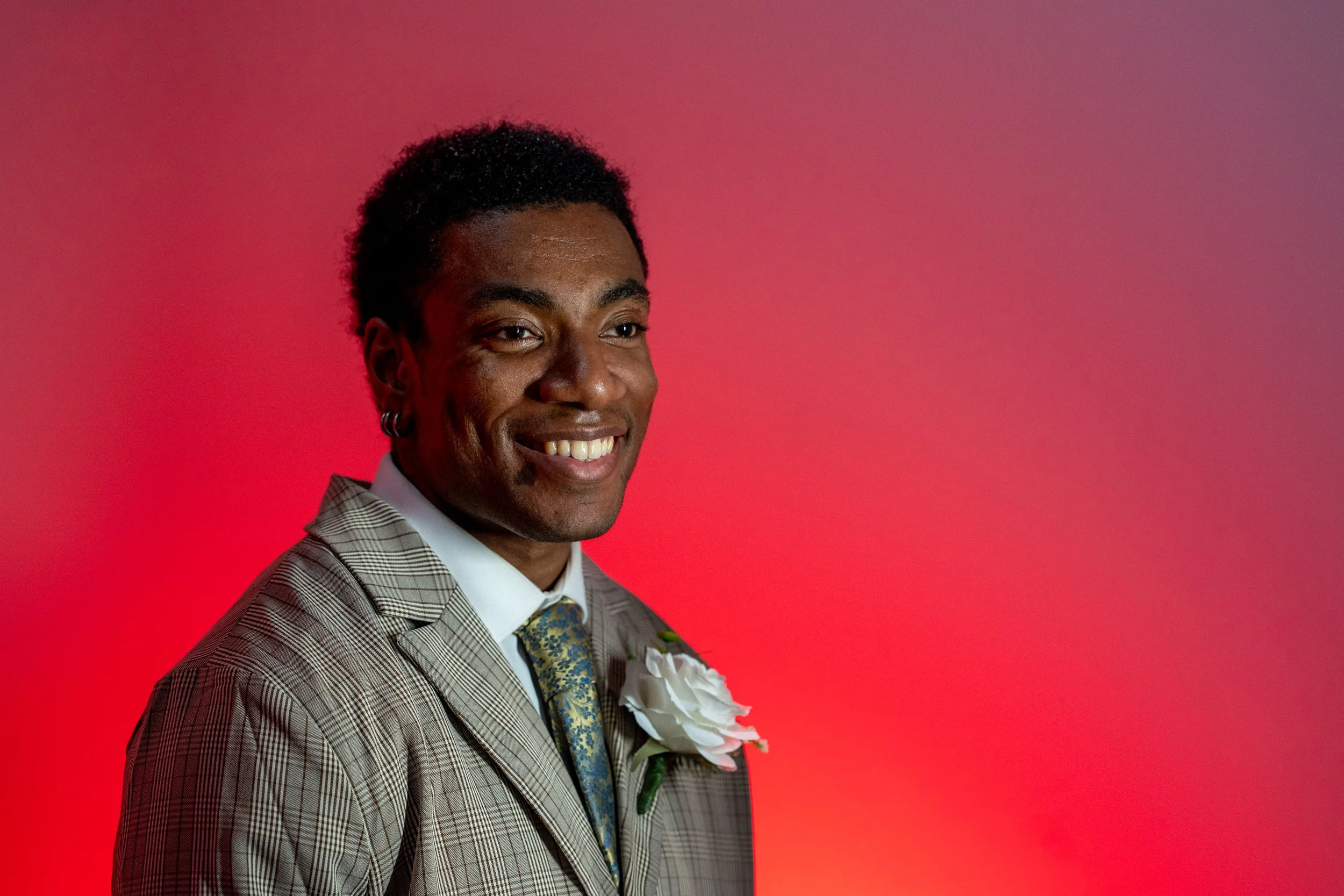 A man in a plaid suit with a floral tie and white shirt, smiling, standing against a pinkish-red background.