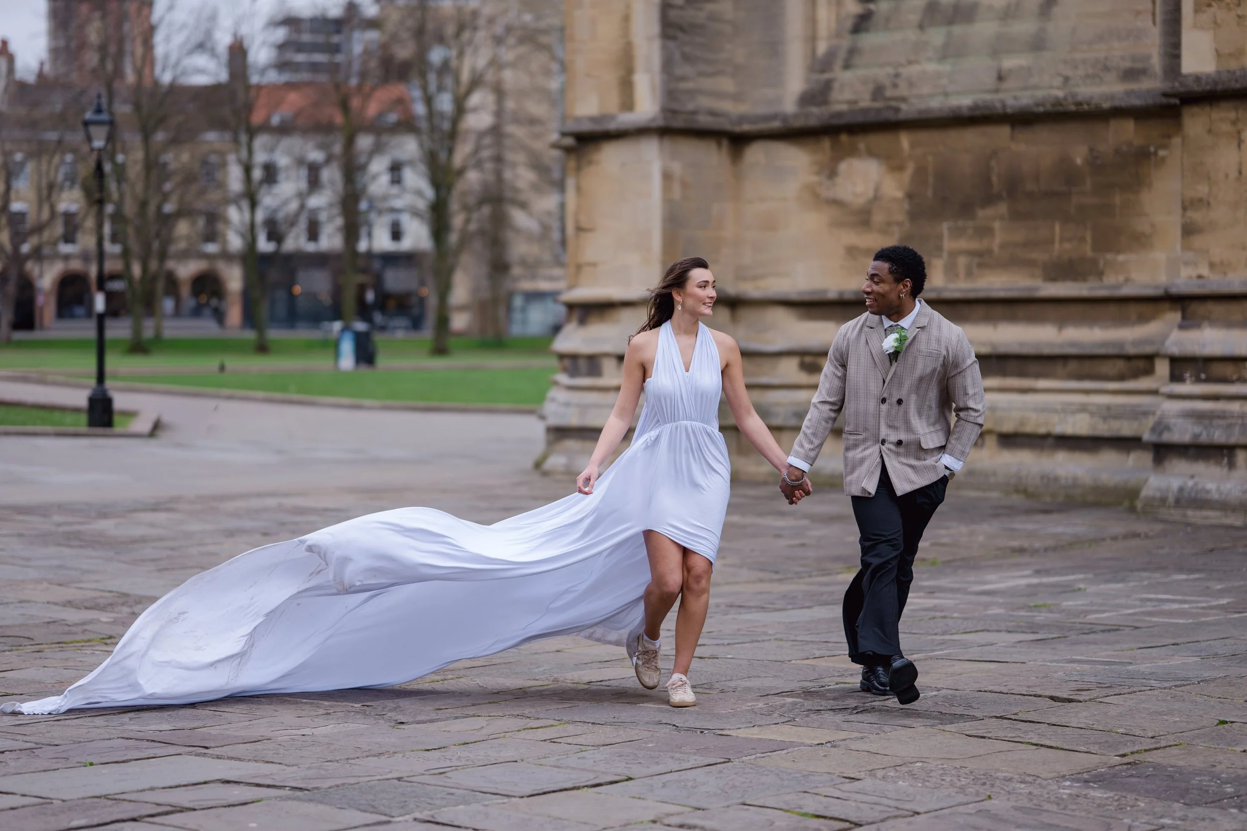 A bride and groom holding hands and walking outside on a stone pavement, with the bride wearing a white wedding dress with a long train and the groom in a light plaid suit with a boutonniere, smiling at each other.