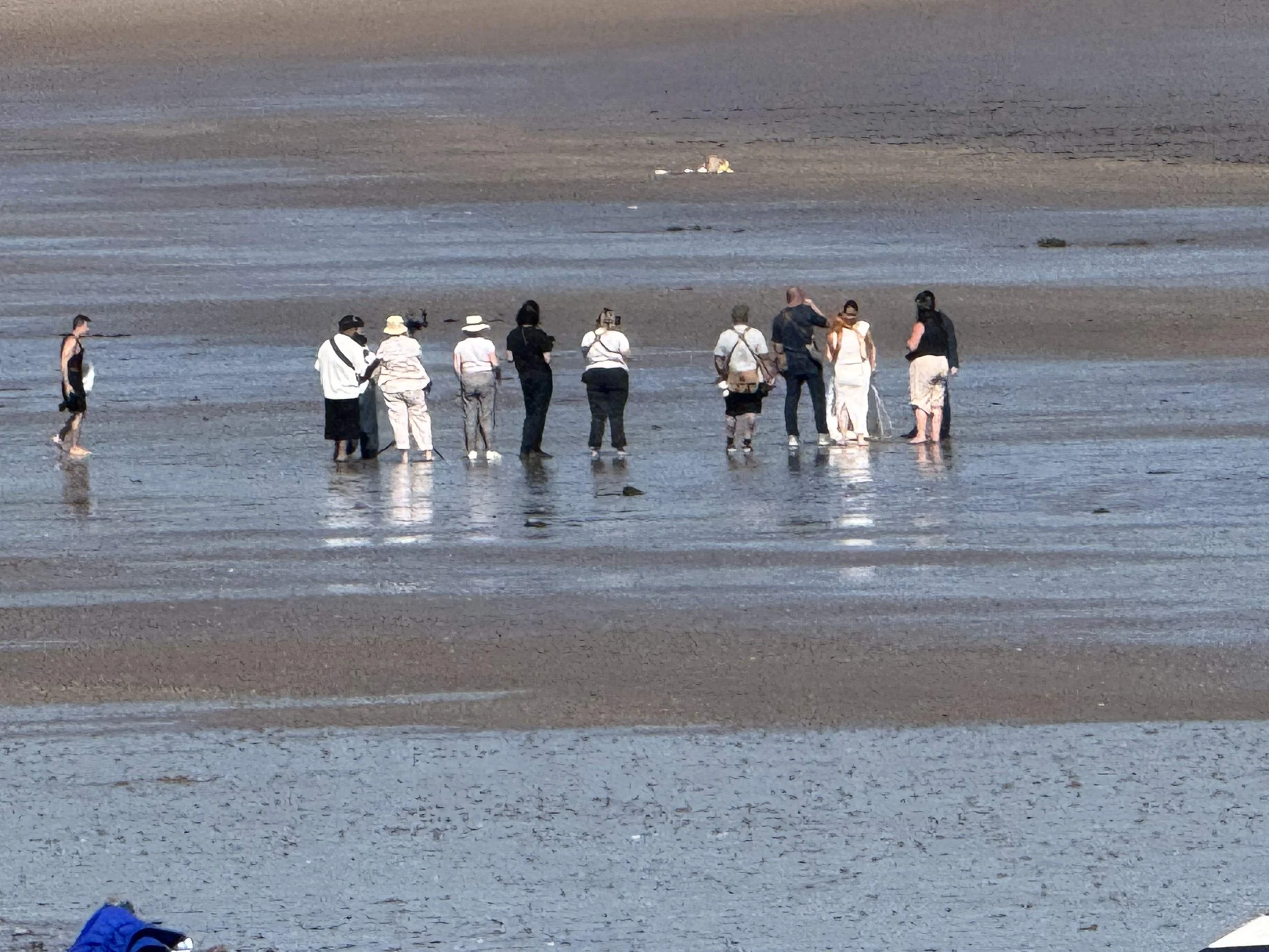 Group of people gathered on a wet, sandy beach, some taking photos and others conversing, with a small dog lying in the distance.