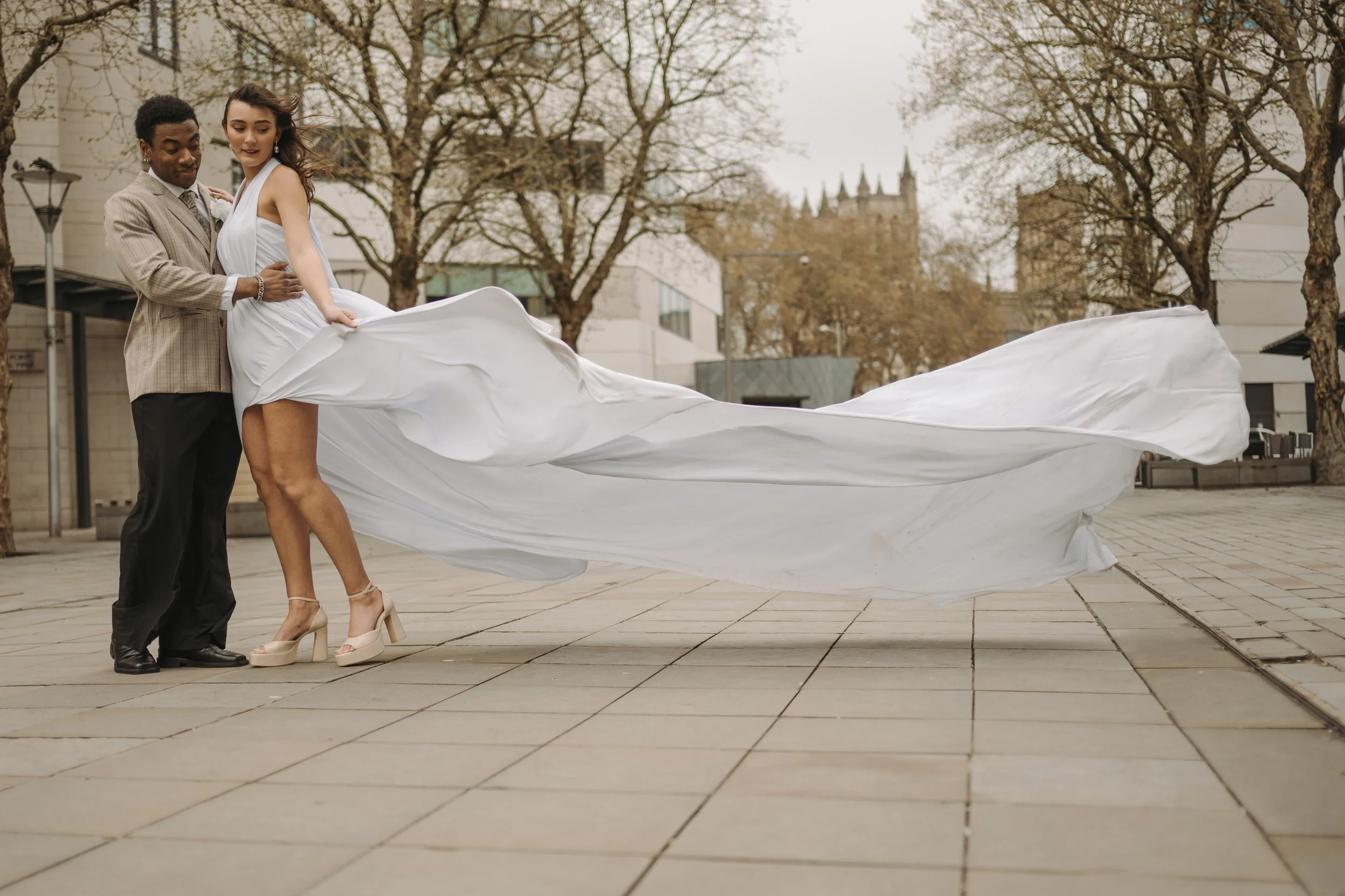 A couple dancing outdoors, with the woman in a white dress and the man in a beige suit, while holding her dress's train. Bare trees and a cityscape with a castle-like structure are in the background.
