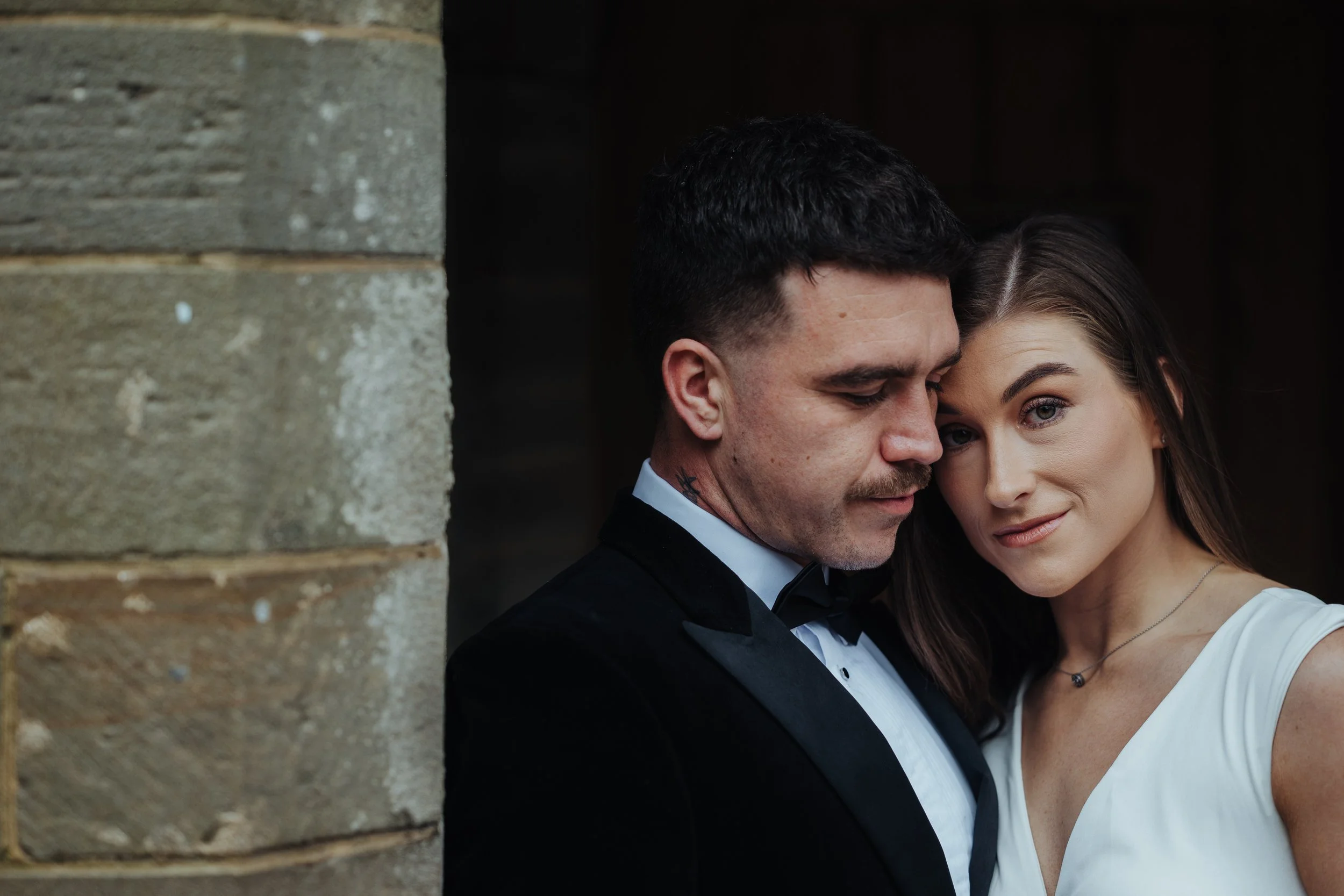 A couple dressed in formal attire, the man in a black tuxedo and the woman in a white dress, close together and smiling softly, standing outside near a stone wall.