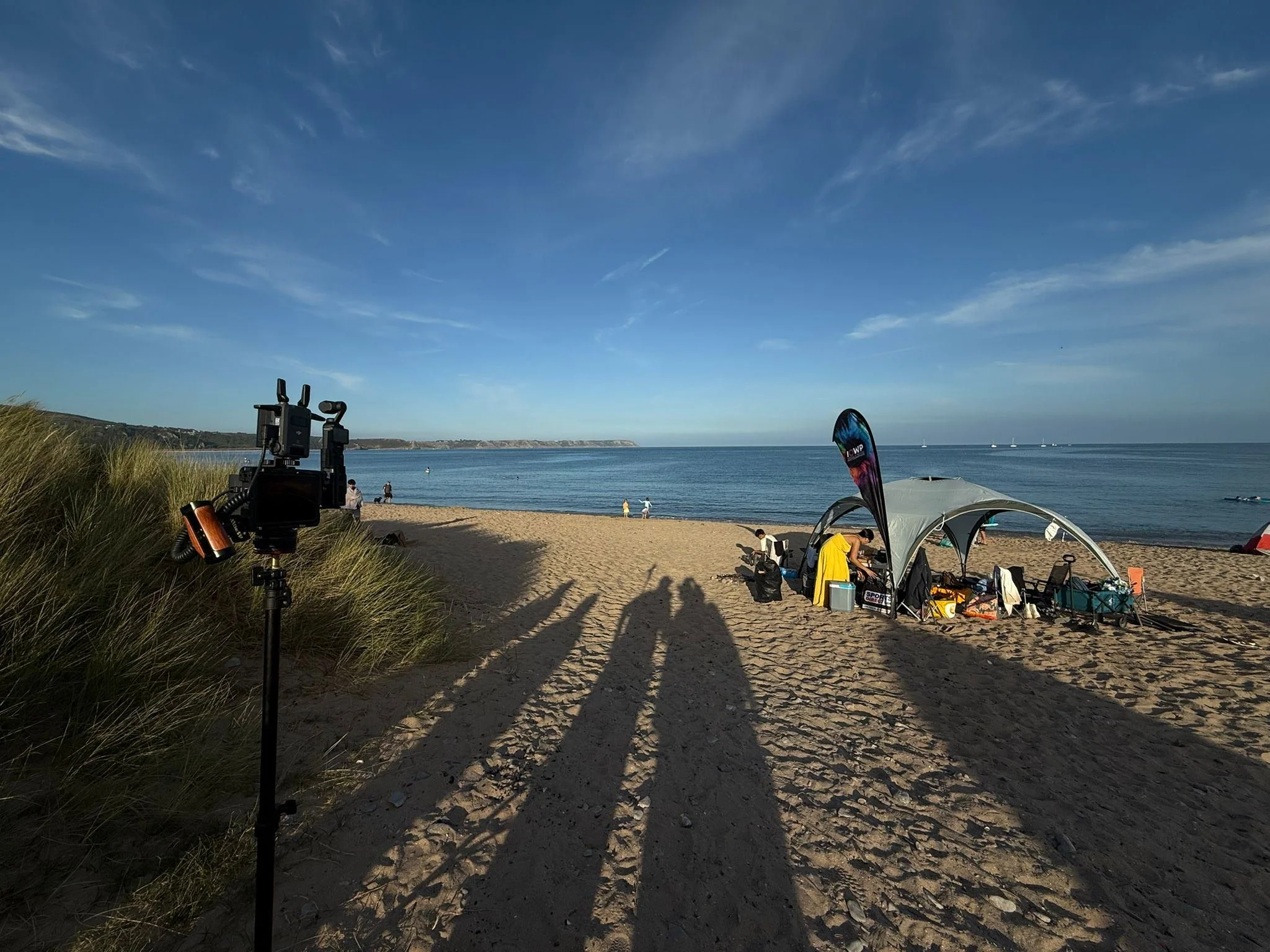Beach scene with a camera on a tripod near grassy sand dunes, a small tent and flag, and people near the shoreline under a blue sky with scattered clouds.