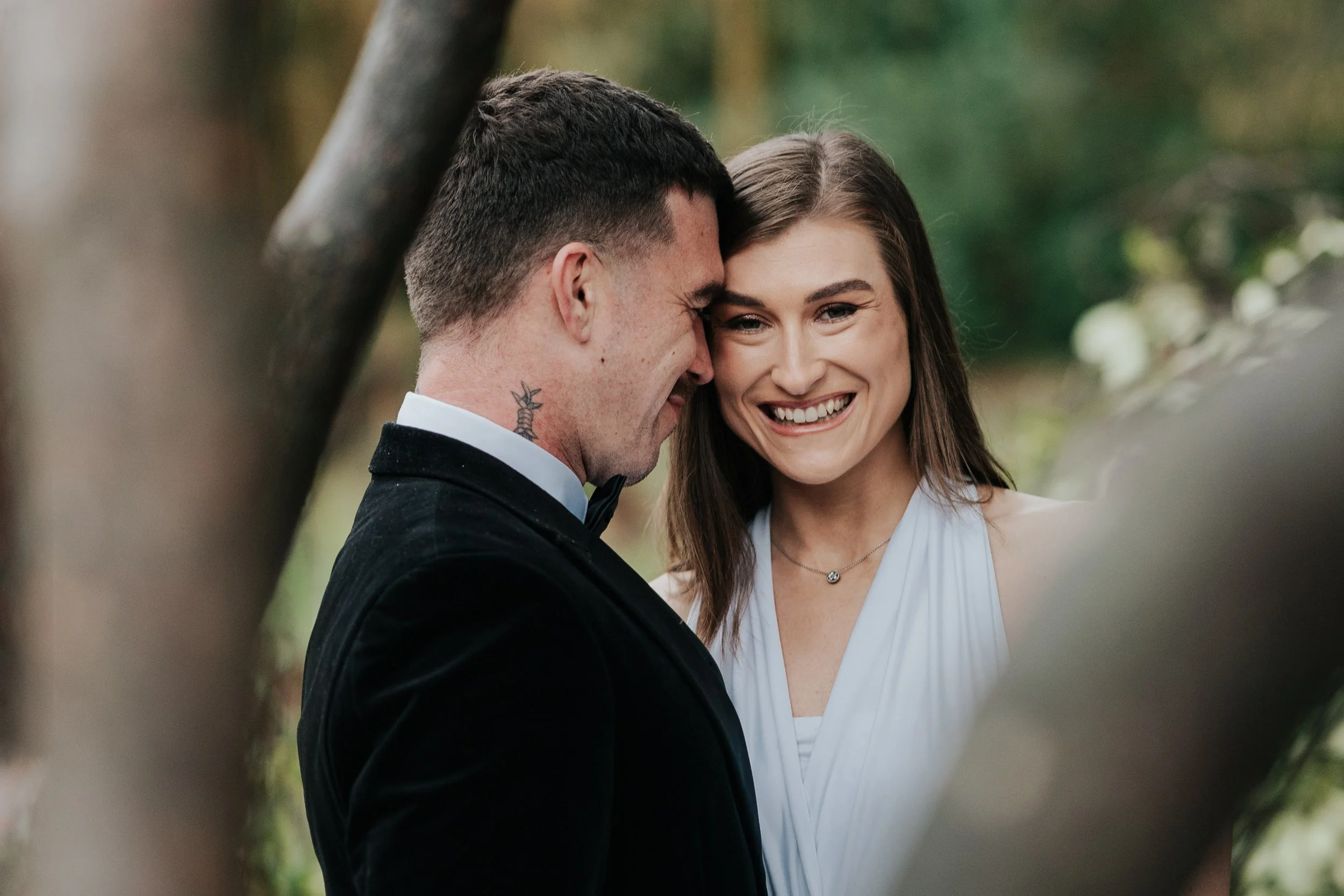A man and woman are smiling closely together outdoors, with their foreheads touching. The man is wearing a black tuxedo, and the woman is in a white dress. They appear to be celebrating a special moment.