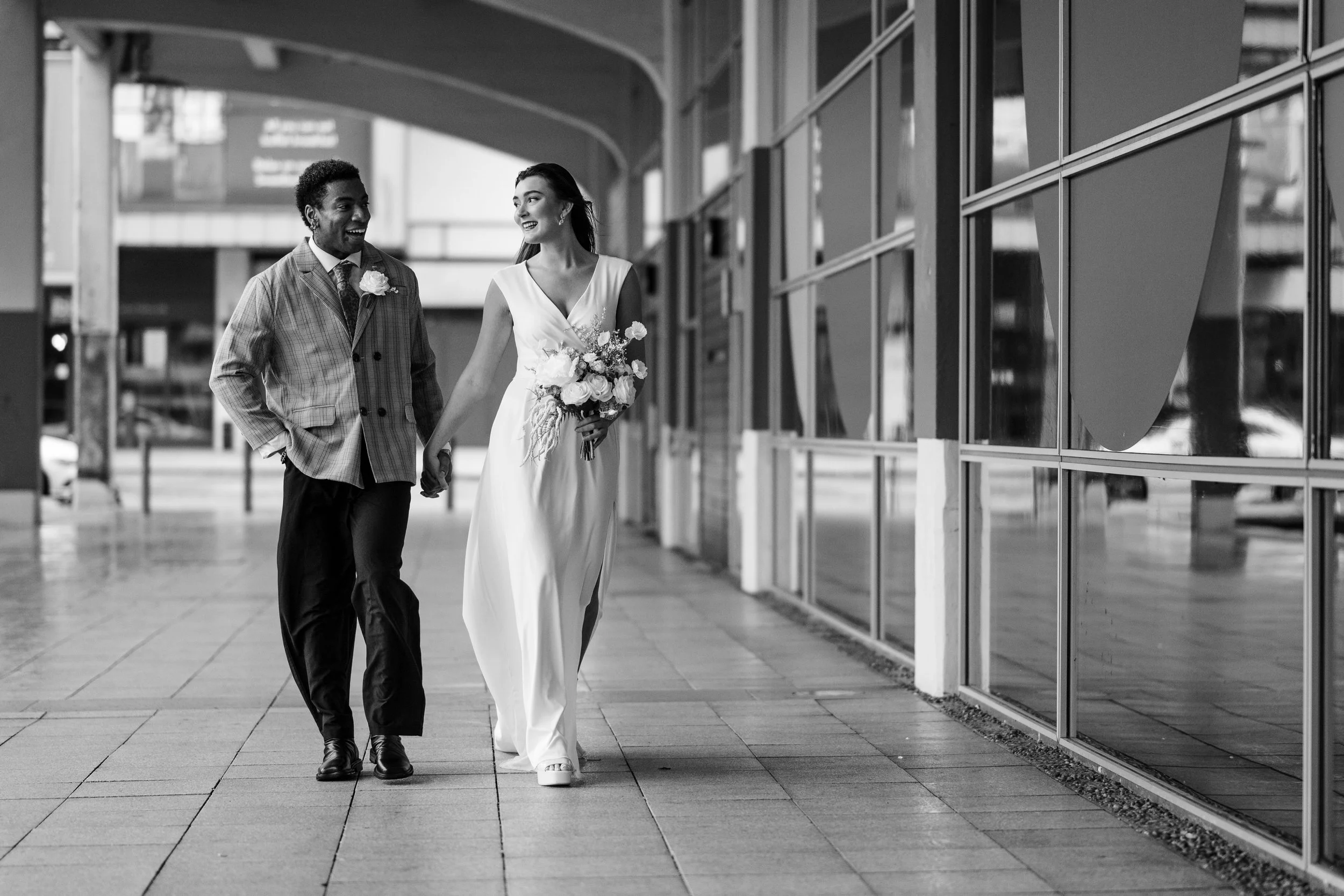 Black and white photo of a happy couple walking hand-in-hand outside a building, with the woman holding a bouquet of flowers. The man is dressed in a plaid suit, and the woman is wearing a white dress, suggesting a wedding or special occasion.