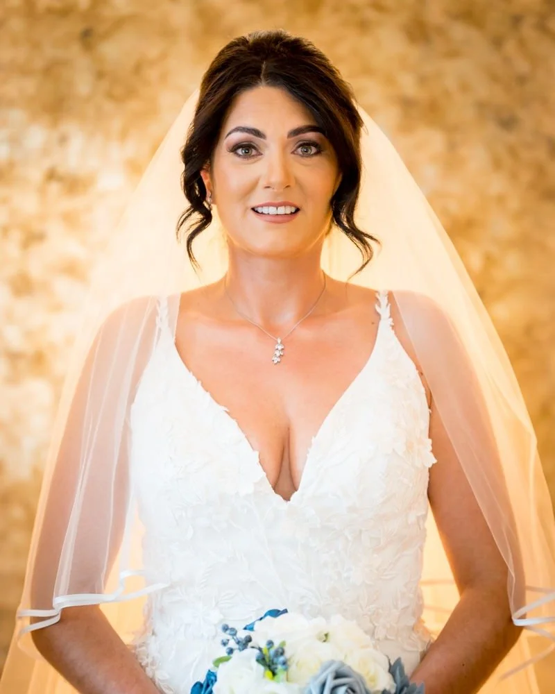 A bride in a white wedding dress holding a bouquet of white and gray roses, standing indoors with a warm, golden background.
