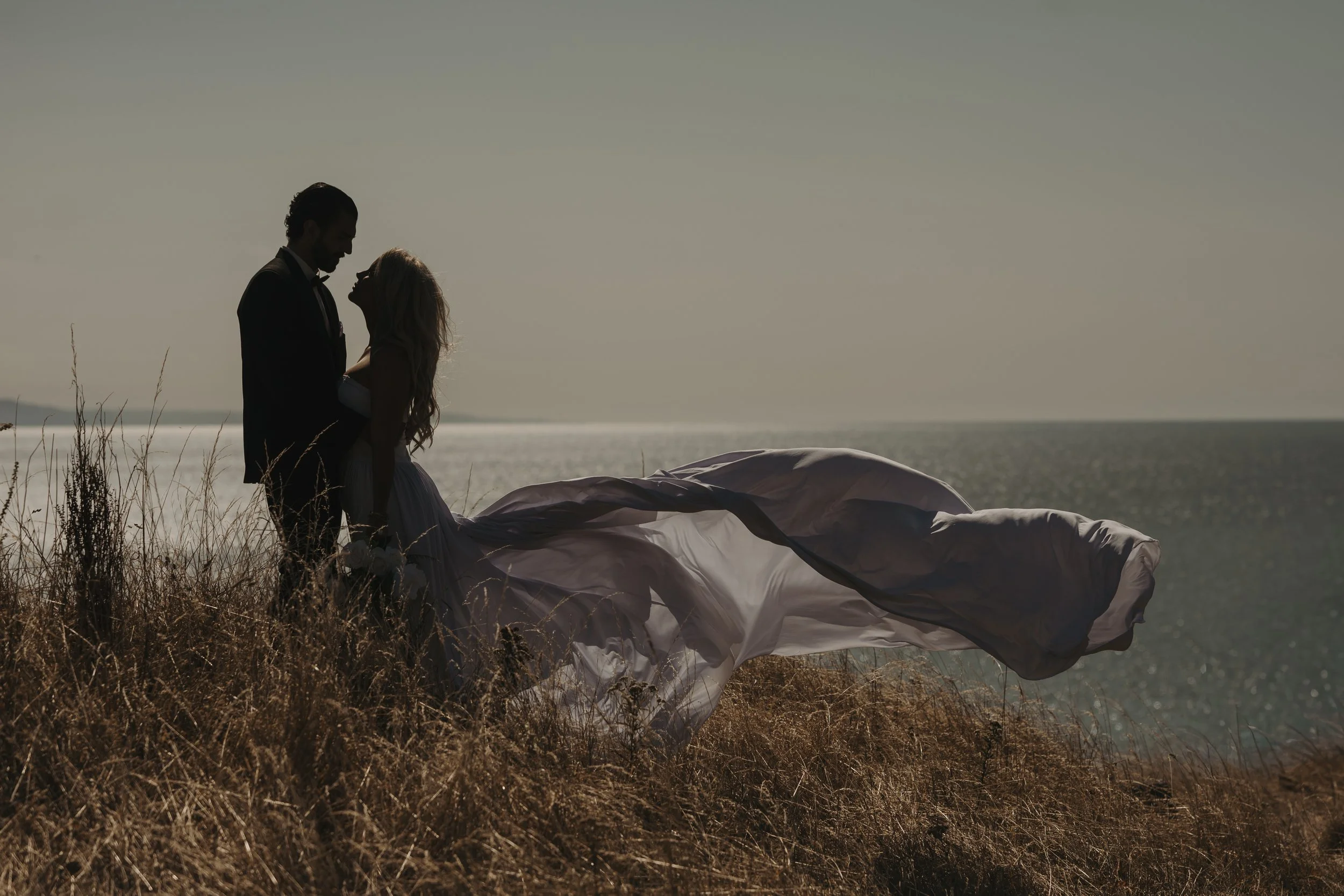 Silhouette of a couple in formal attire standing close on a grassy cliff overlooking the ocean at sunset, with a wedding dress flowing on the ground.
