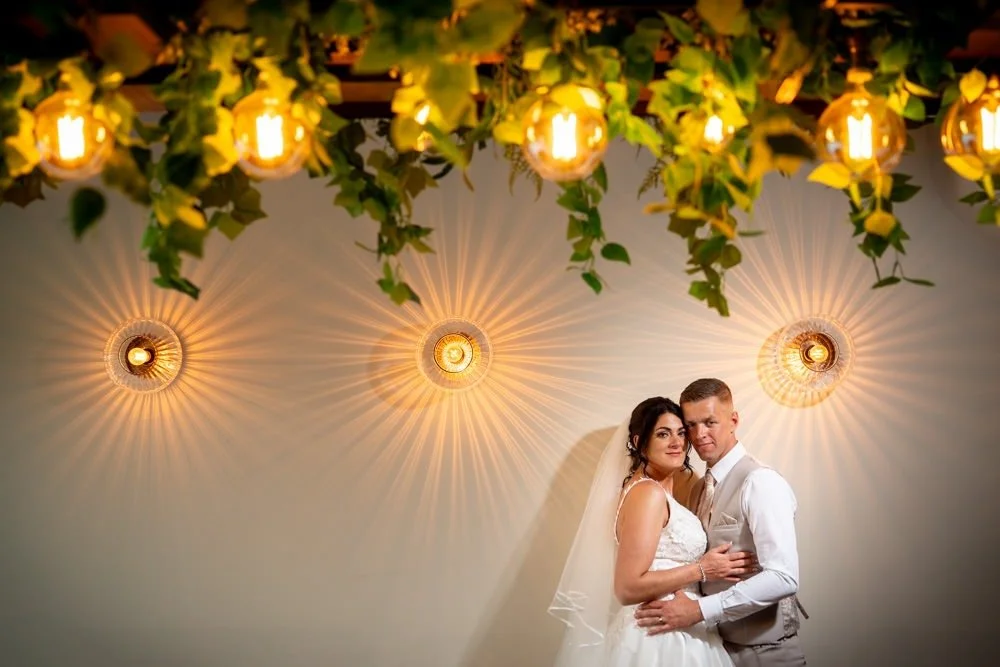 A bride and groom posing together at their wedding, with decorative lighting and greenery overhead.