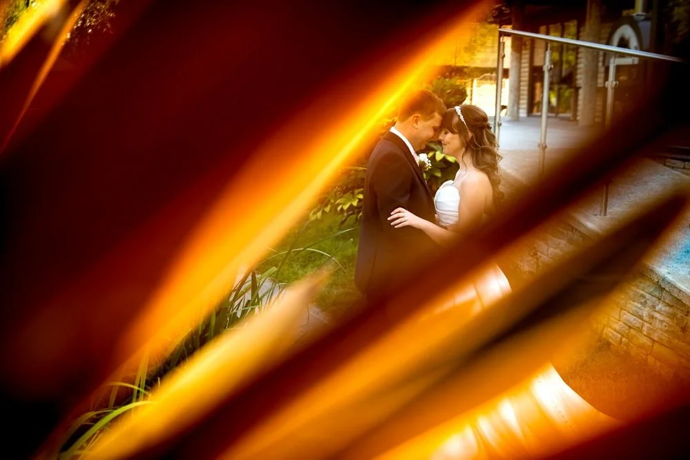 A bride and groom standing close together outdoors, viewed through a partially opened large book or an object with warm lighting, during a wedding ceremony or romantic moment.