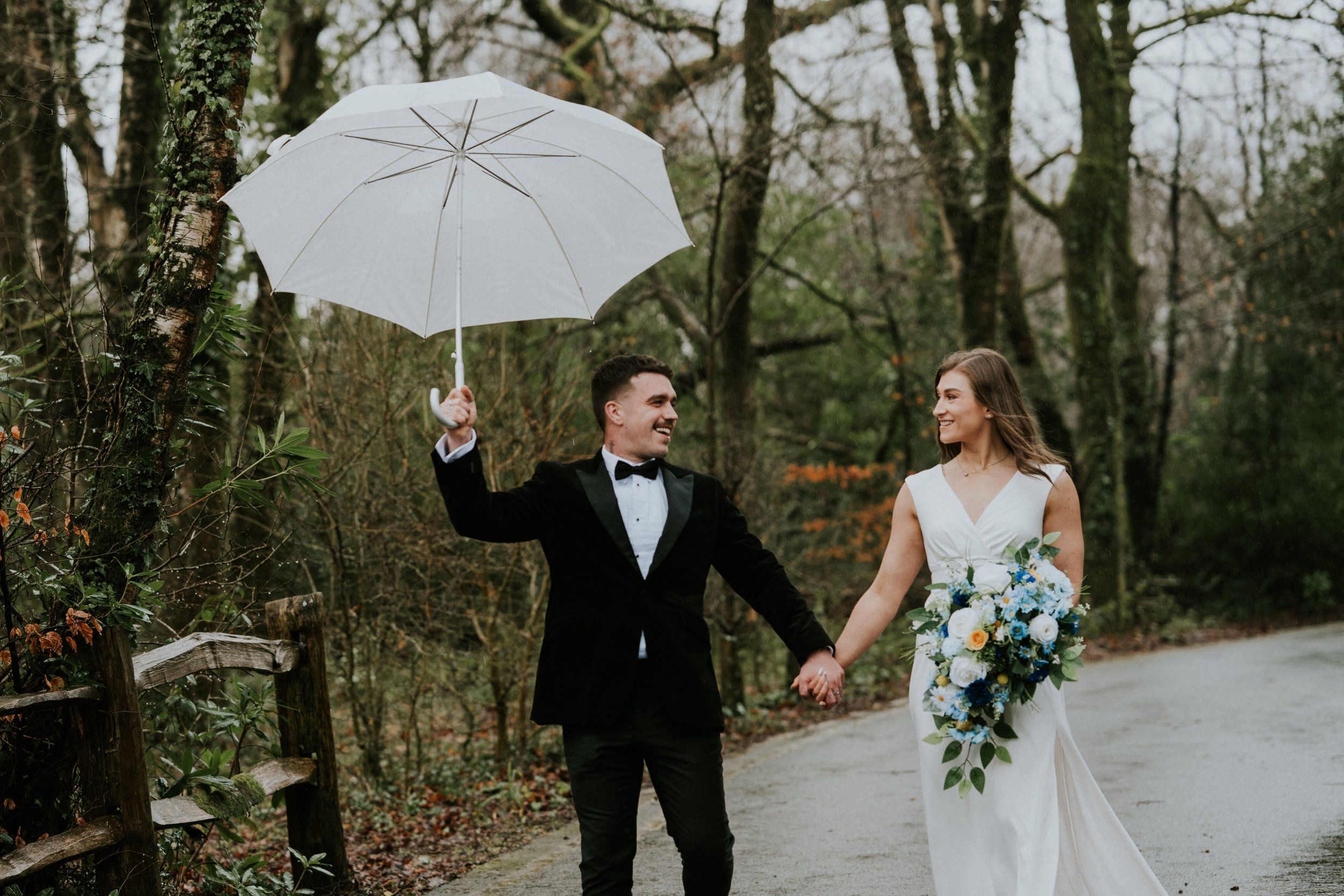 Happy bride and groom holding hands while walking in a forest, the groom holding a white umbrella, on a rainy day.
