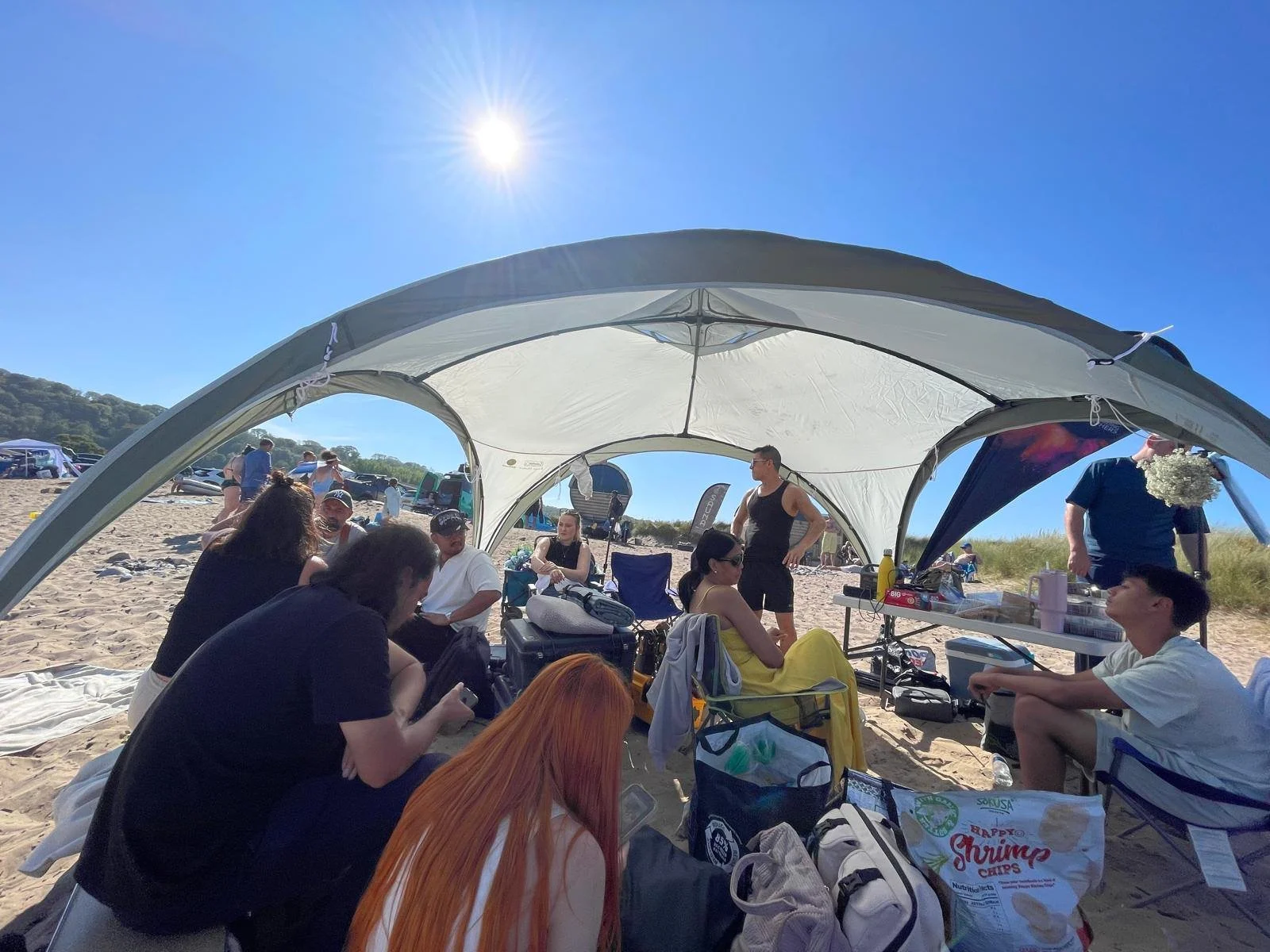 People sitting and standing under a large beach tent on a sandy beach, with items like bags, drinks, and snacks nearby, and others in the background near parked cars and dunes under a sunny, clear blue sky.