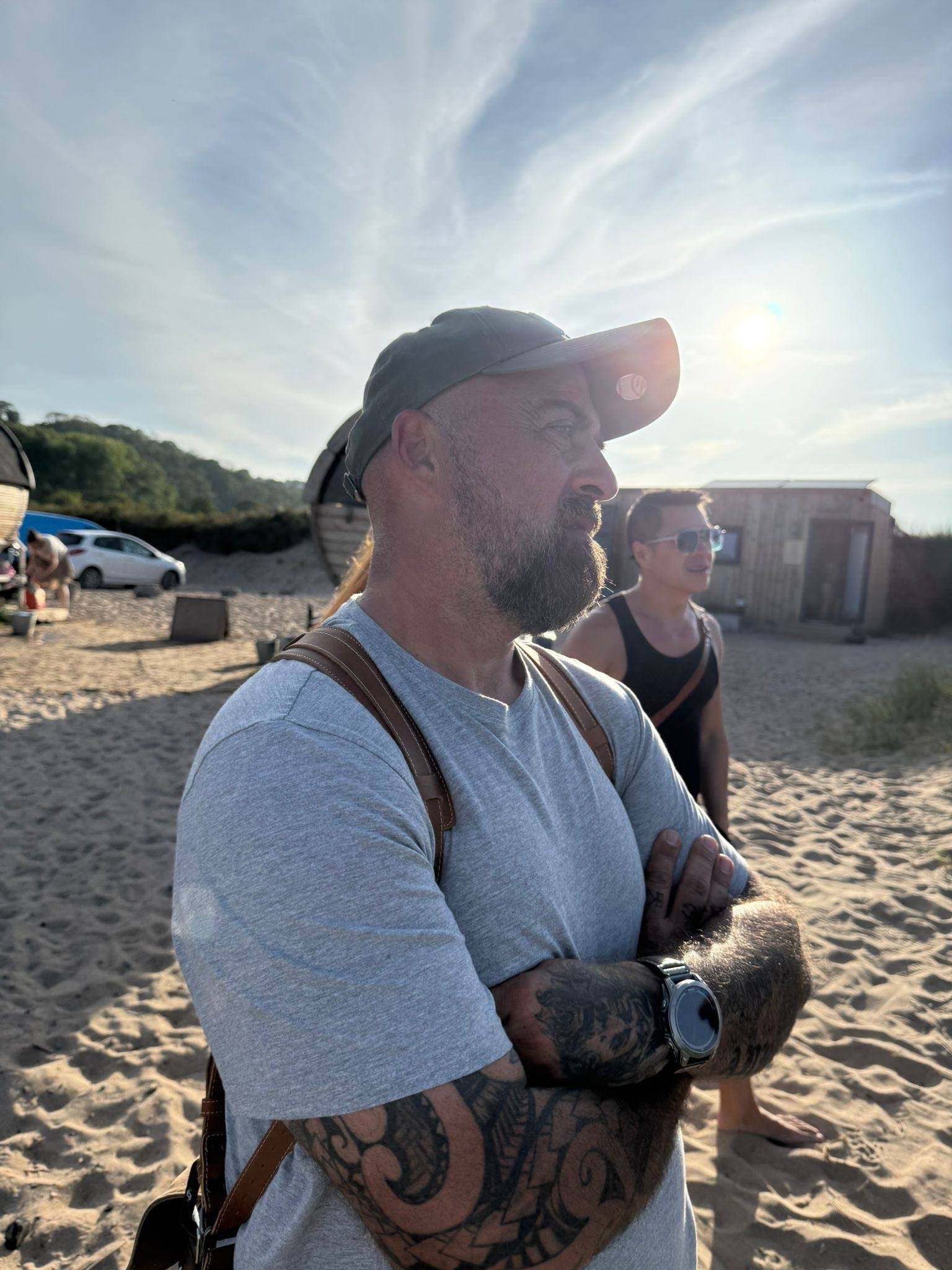A man with a beard and tattoos on his arms standing on a sandy beach with crossed arms, wearing a gray T-shirt, cap, and a watch, with another person in the background and a building nearby, bathed in sunlight.