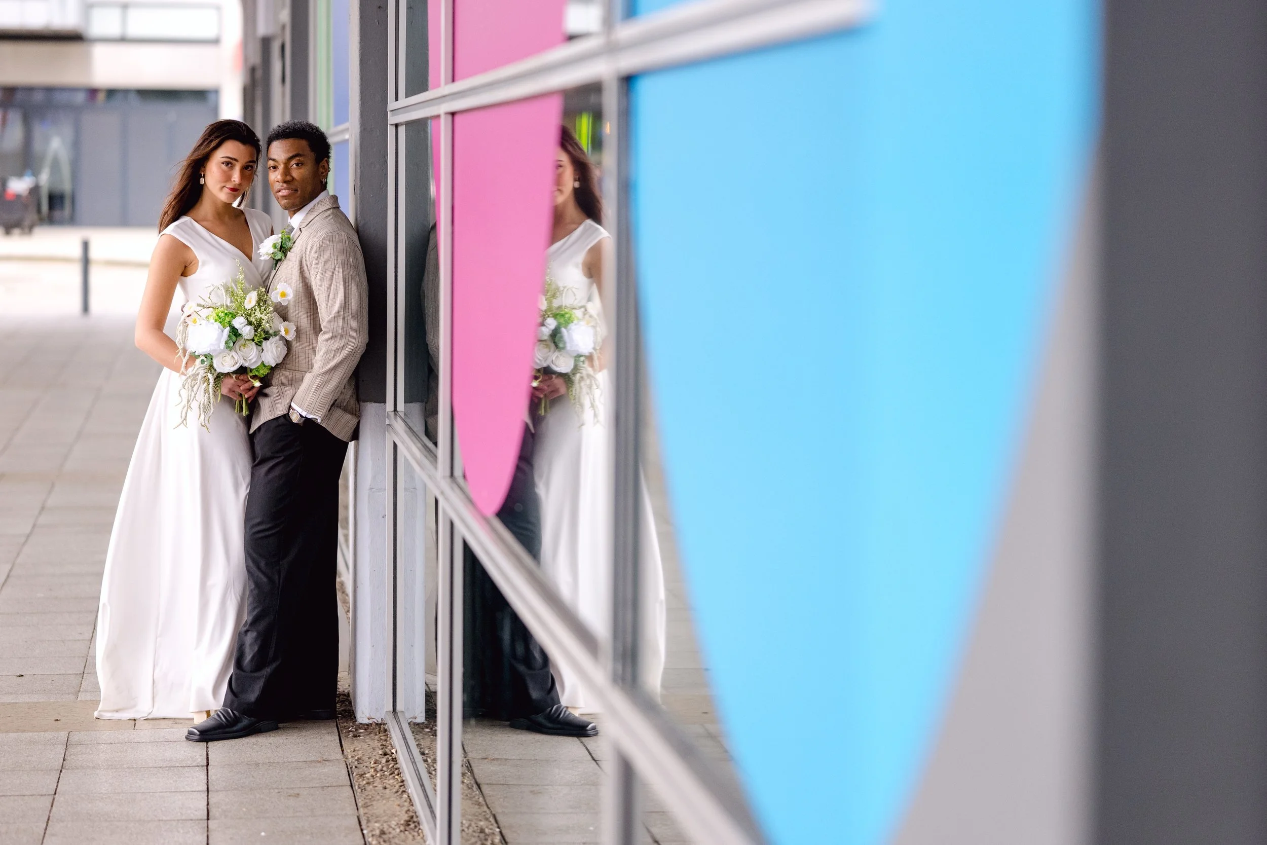 A bride and groom standing close together outside near a glass building with colorful panels, with their reflections visible on the glass.