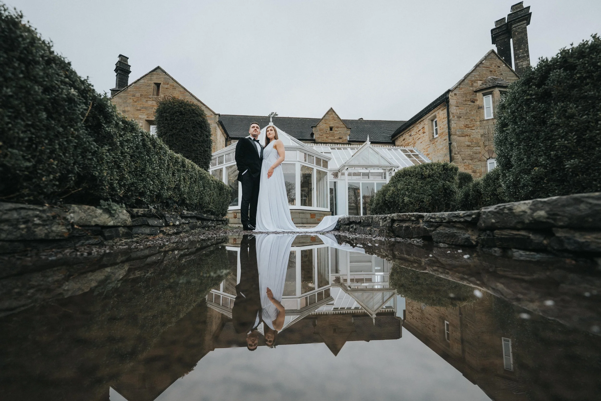 A bride and groom standing outside a stone house with a glass conservatory, reflected in a puddle on the ground.
