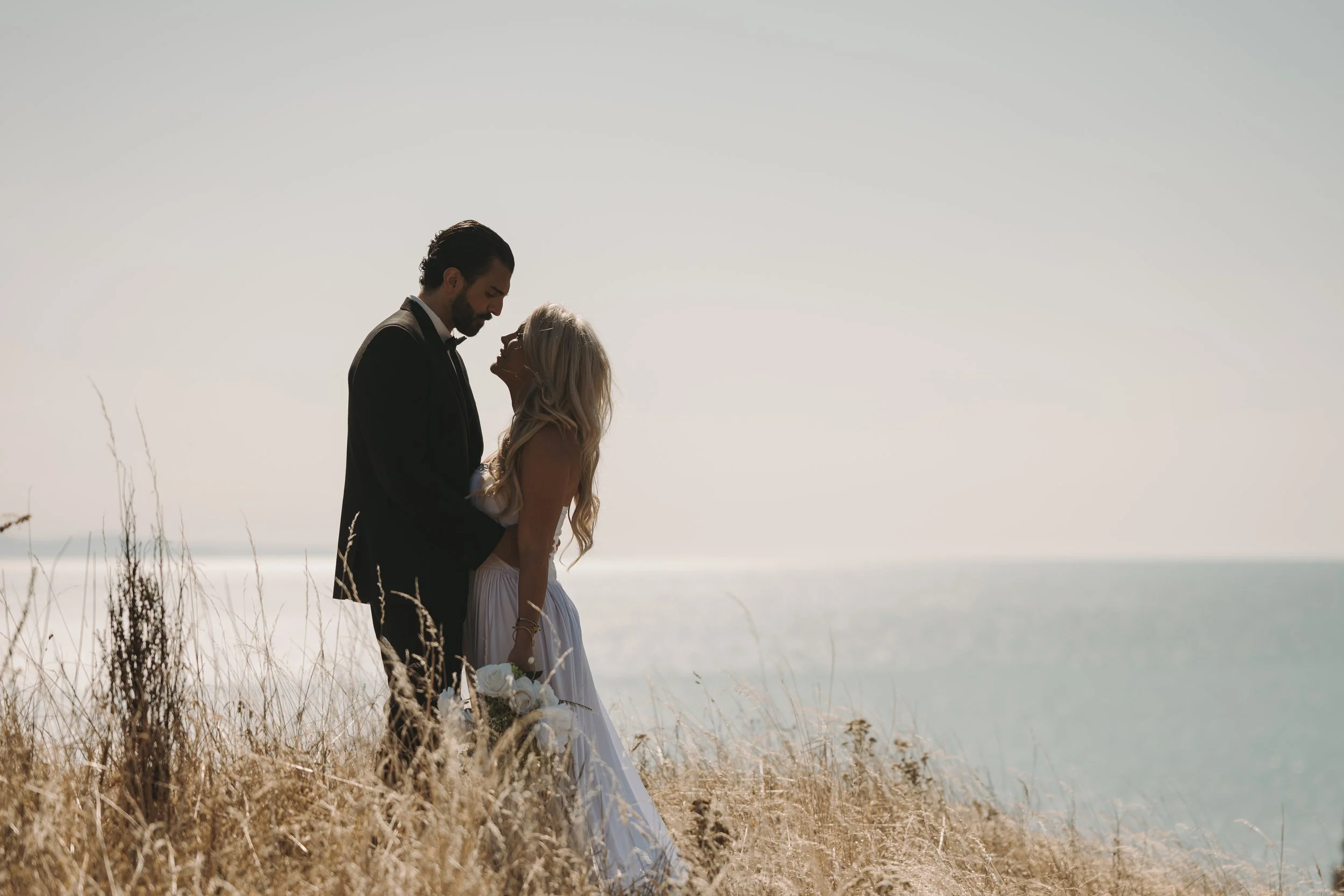 A man and a woman in wedding attire standing close together outdoors during daytime, with the man gently holding the woman and both looking at each other. The background features a clear sky and distant ocean or lake.