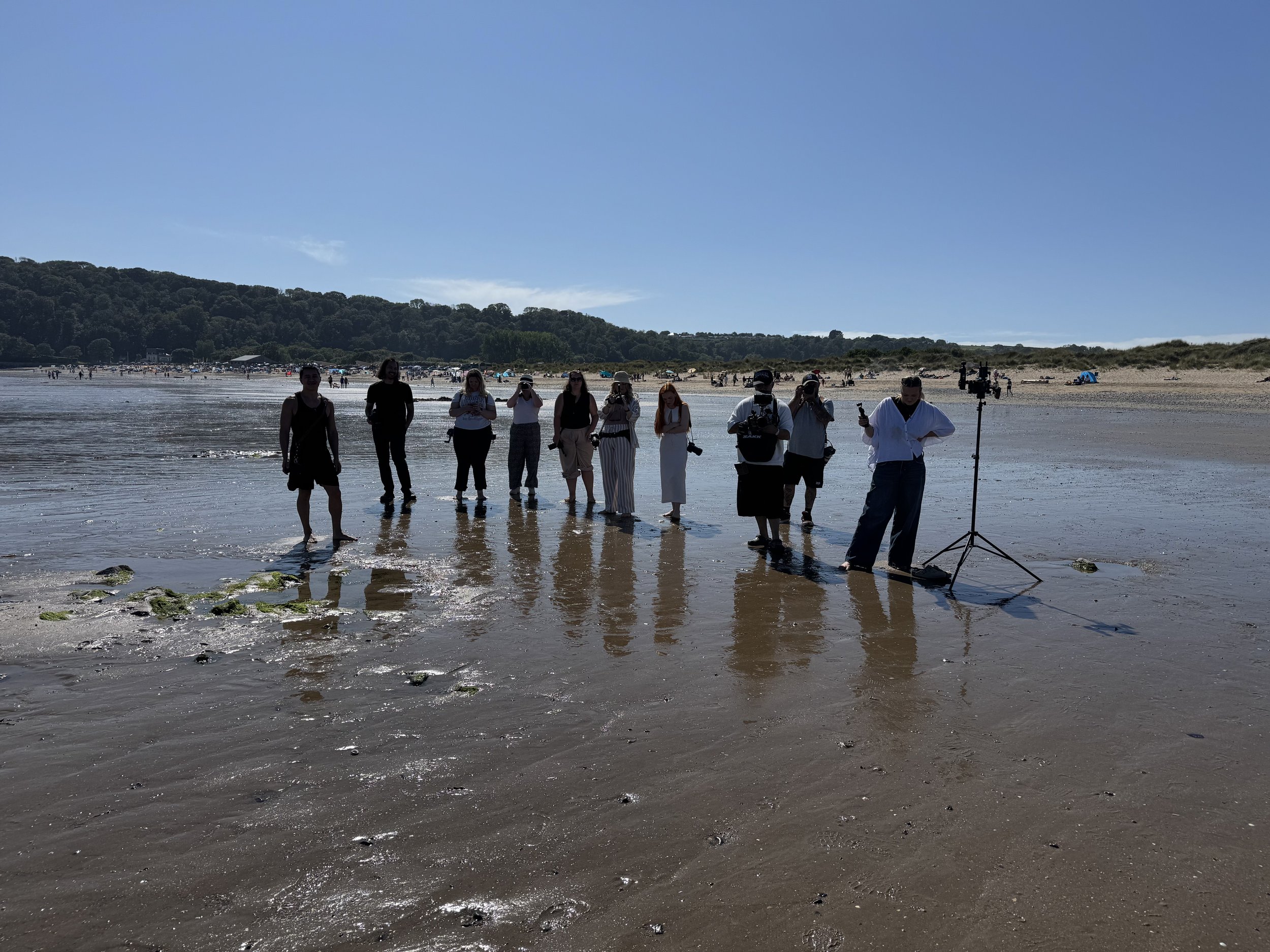 A group of people standing on a wet beach, some taking photos or videos, with one person operating filming equipment, under a bright blue sky with distant sand dunes and a hillside covered in trees.