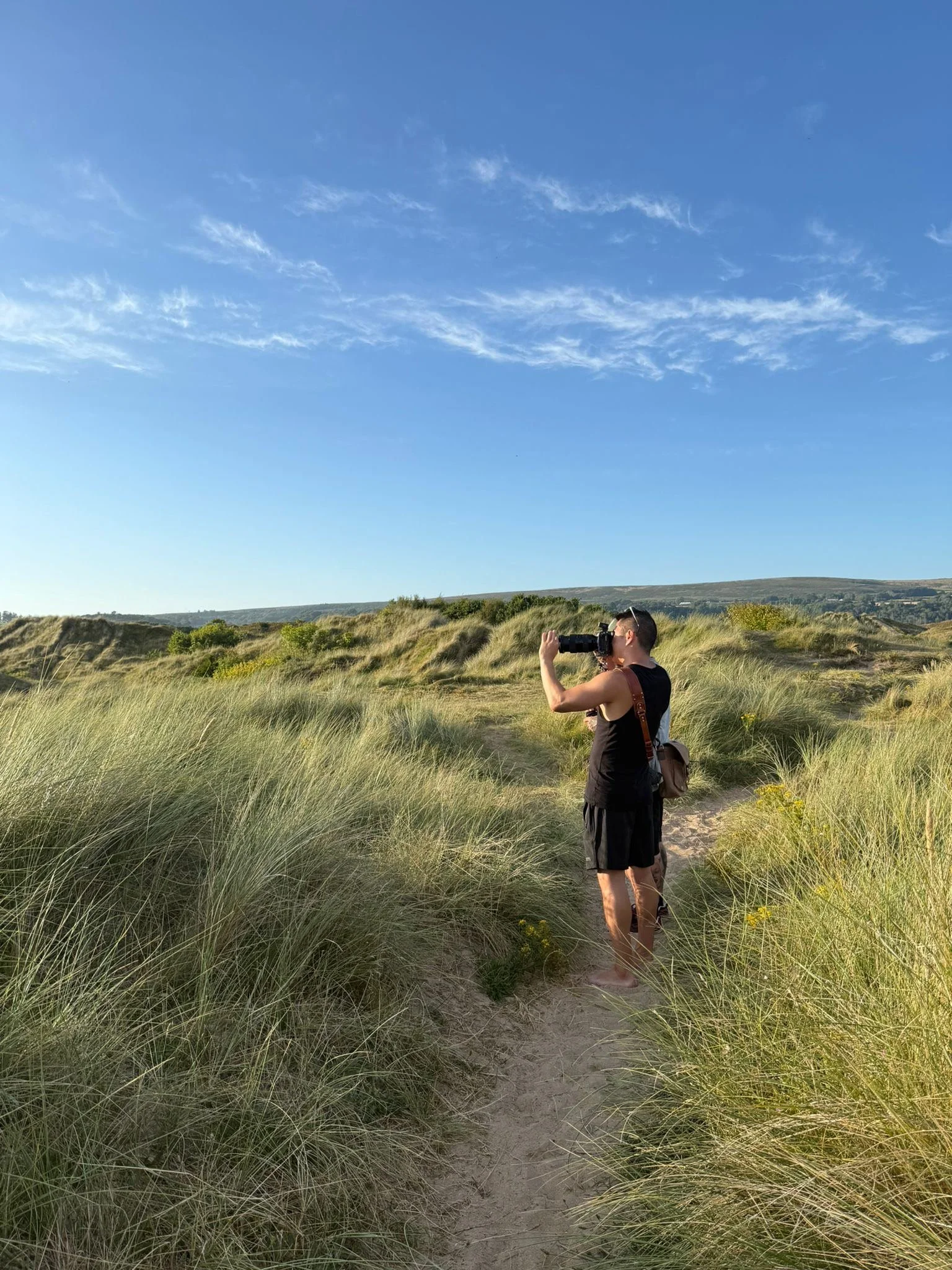 A person standing on a sandy path in tall grass, looking through binoculars in a natural outdoor setting with a blue sky and some clouds.