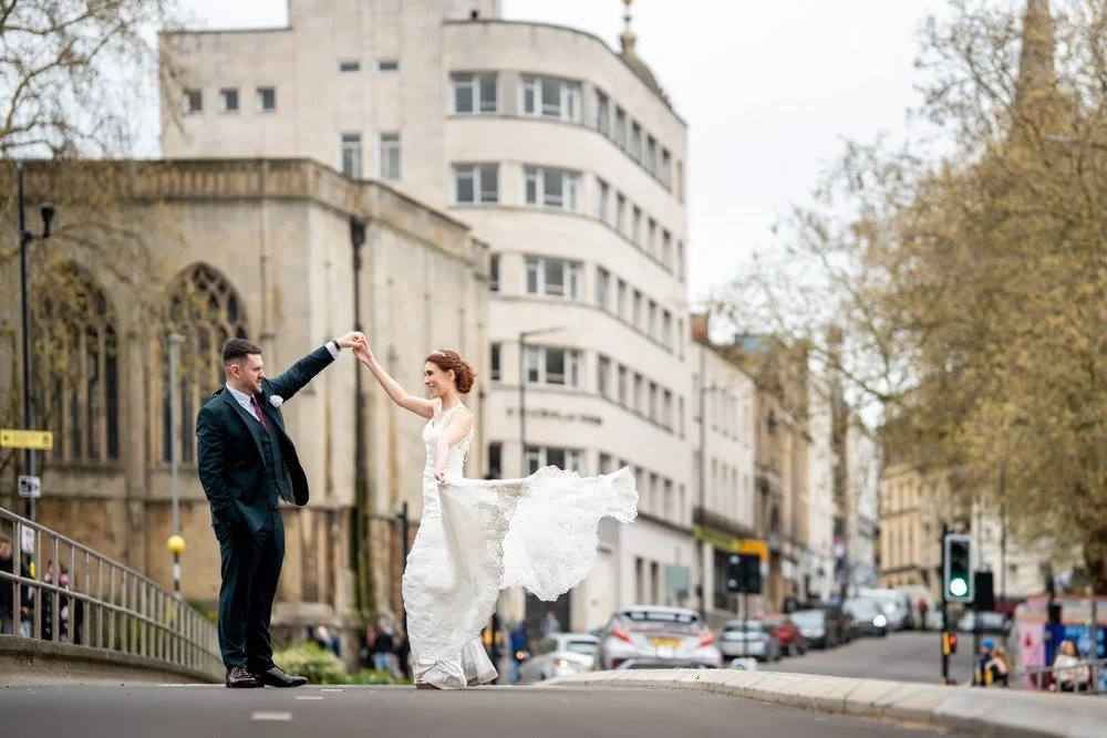 A bride and groom dancing on a city street with buildings and trees in the background.