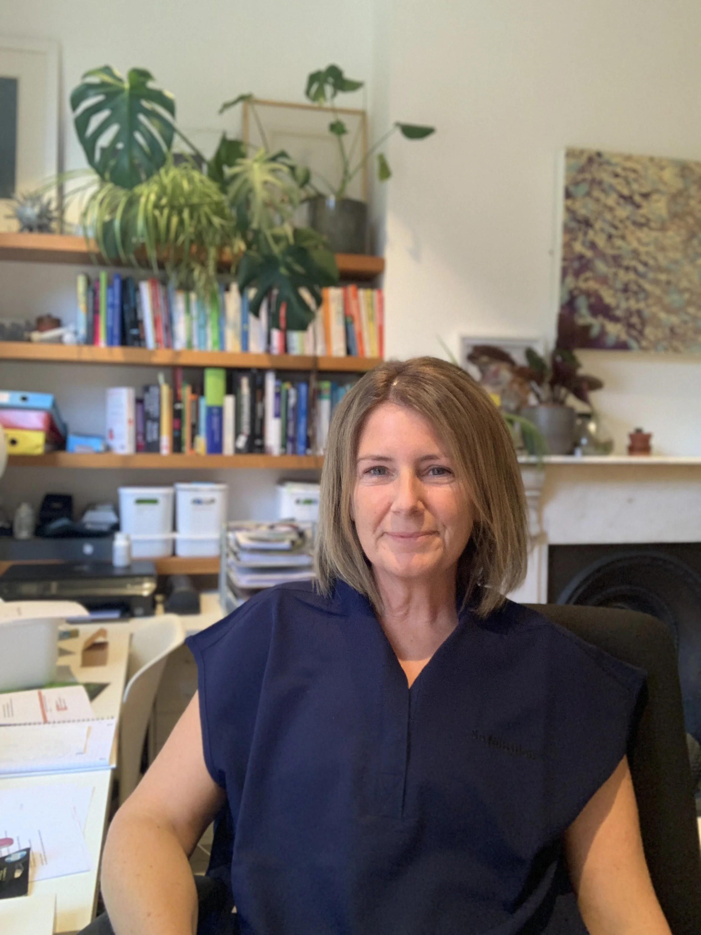 Jayne Higgins, a middle-aged woman with shoulder-length brown hair and green eyes, wearing a dark blue shirt, sitting indoors with a bookshelf and plants in the background.