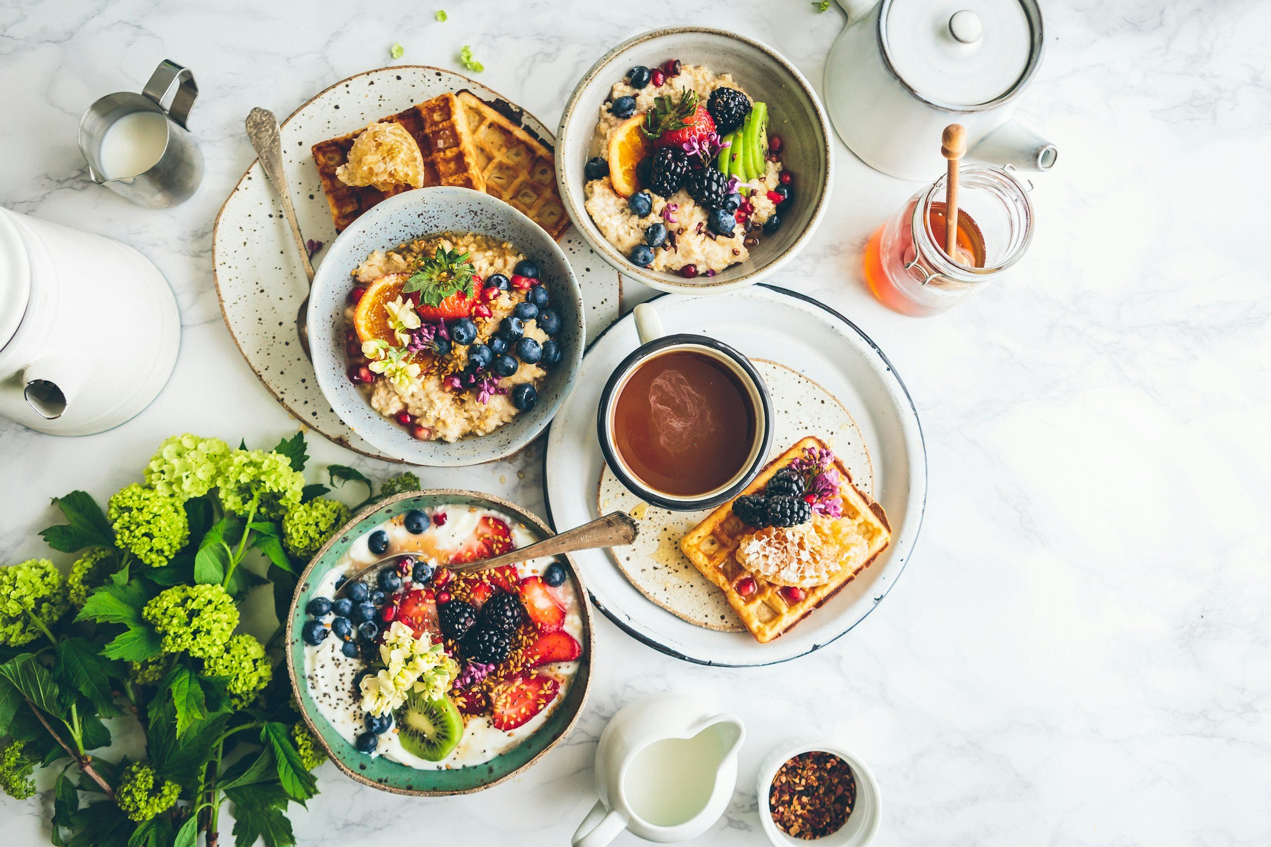 Assorted breakfast items including waffles, oatmeal topped with berries and fruit, a bowl of yogurt with berries and flowers, a cup of tea, a glass of milk, and small bowls of honey and crushed red pepper, arranged on a white marble surface with green flowers.