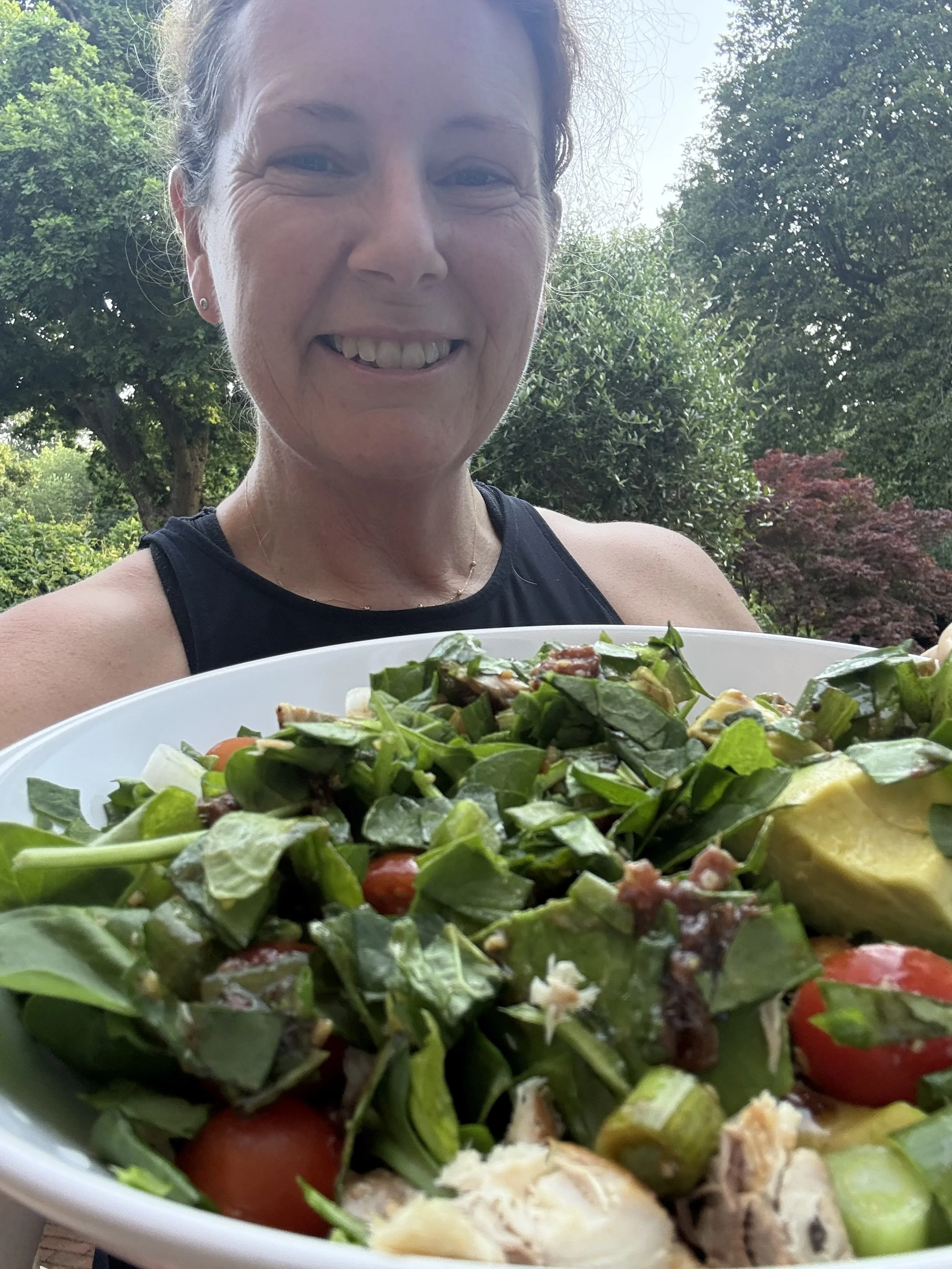 A smiling woman outdoors holding a bowl of salad with greens, cherry tomatoes, and other vegetables, with trees and bushes in the background.