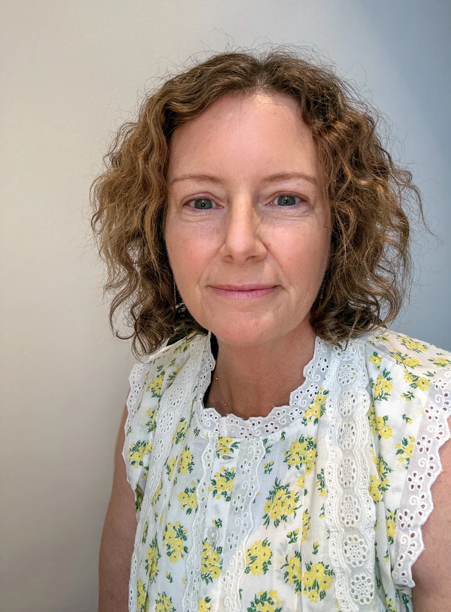 A woman with curly brown hair and blue eyes, wearing a white dress with a yellow floral pattern and eyelet lace trim, standing against a plain wall.